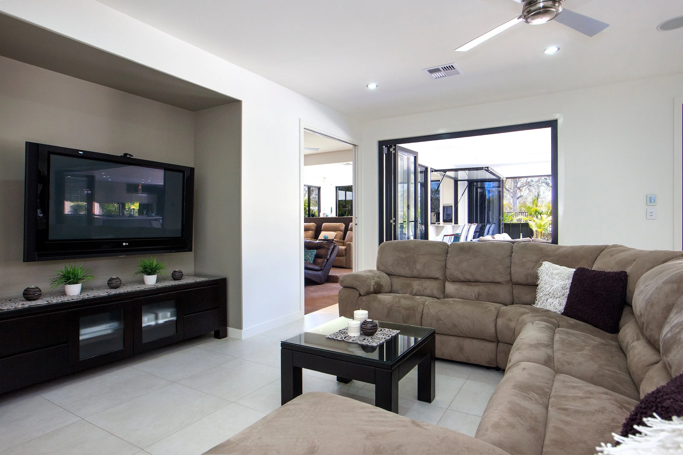 Living room with beige sectional sofa, black coffee table, and black TV stand with small plants and decorative items. Open doorway leads to a sunroom with glass doors and outdoor seating.