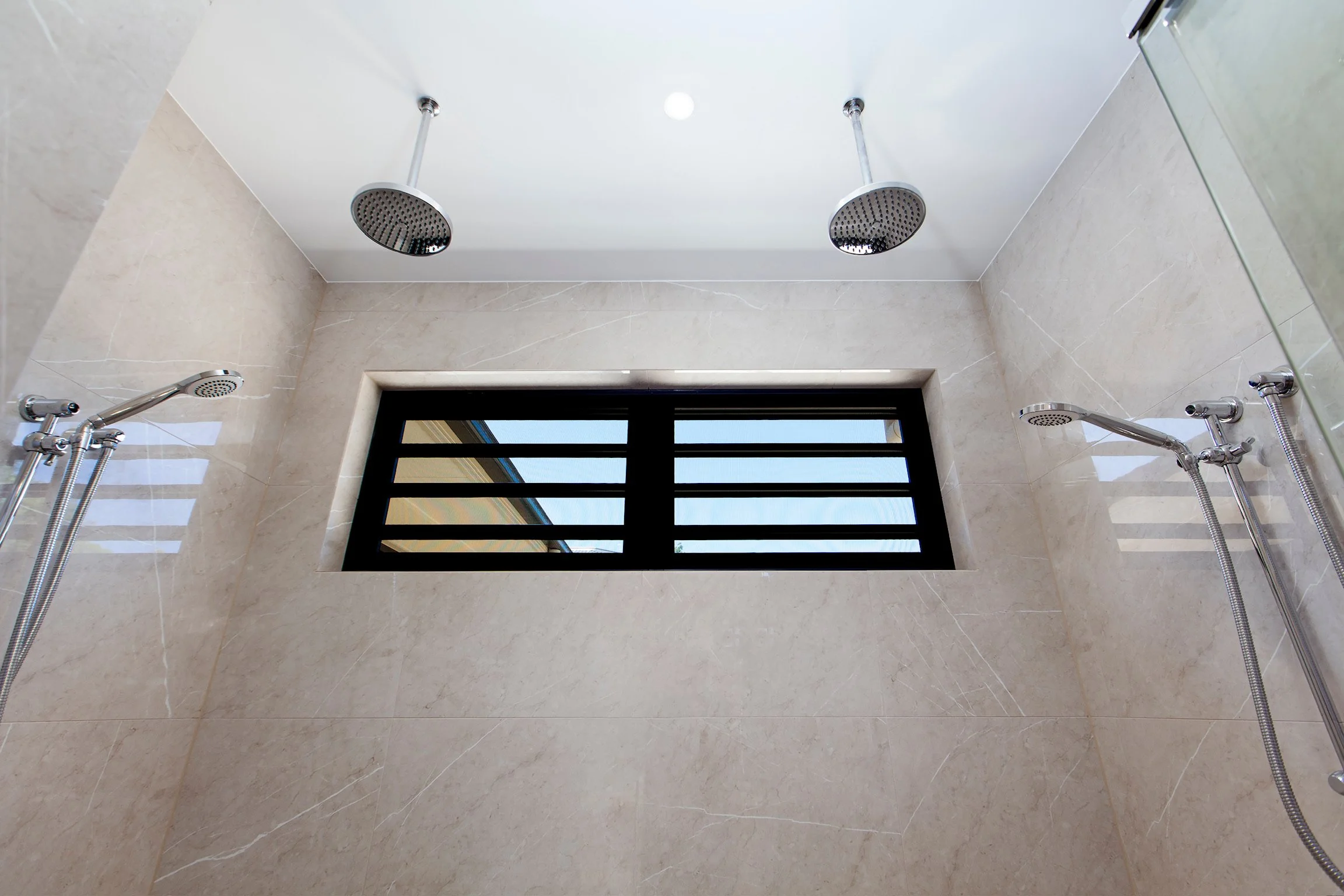 View of a shower ceiling with two round rain shower heads, a window below, and beige tiled walls.