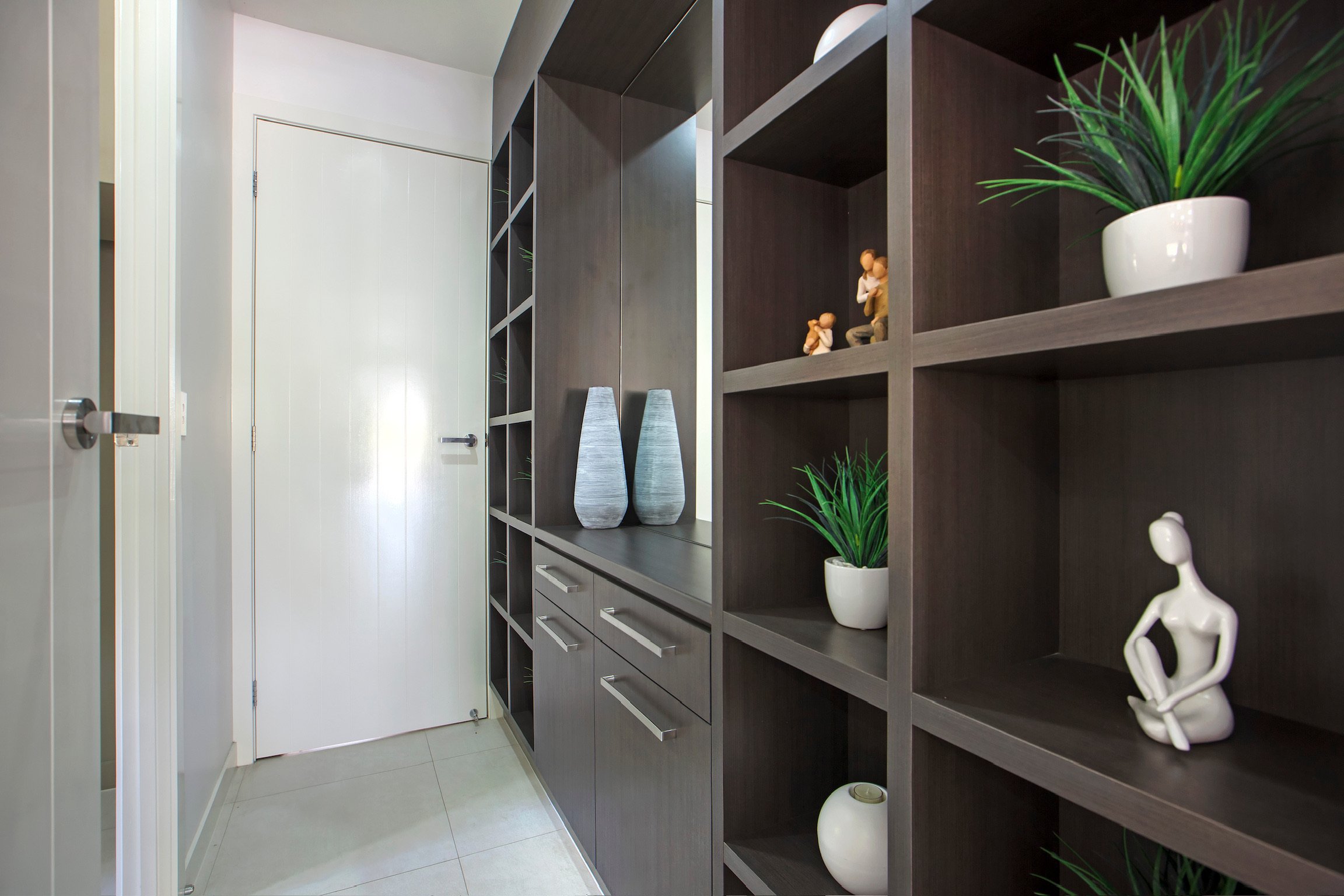 Interior view of a modern room with a dark wooden shelving unit containing decorative plants, vases, and figurines, next to a white door and light-colored tiled floor.