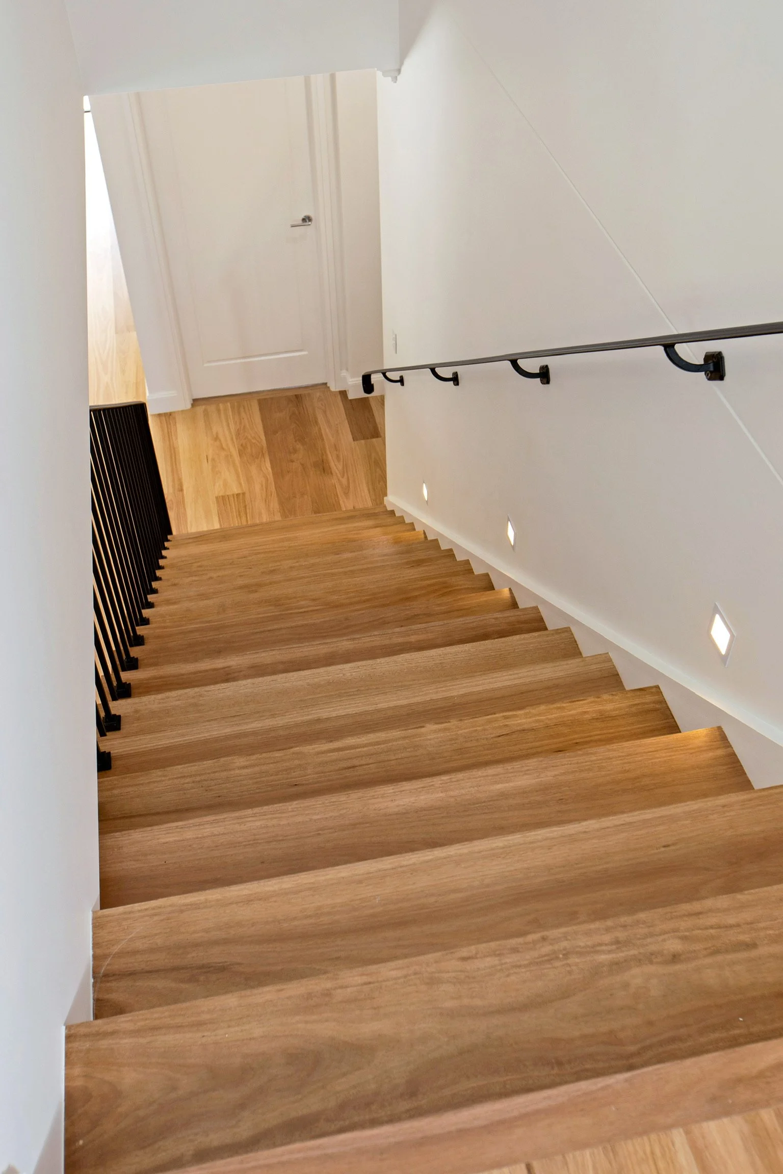 A staircase with wooden steps and black railing, leading down to a small landing. The landing has a white door and wood flooring, with recessed lighting on the wall.