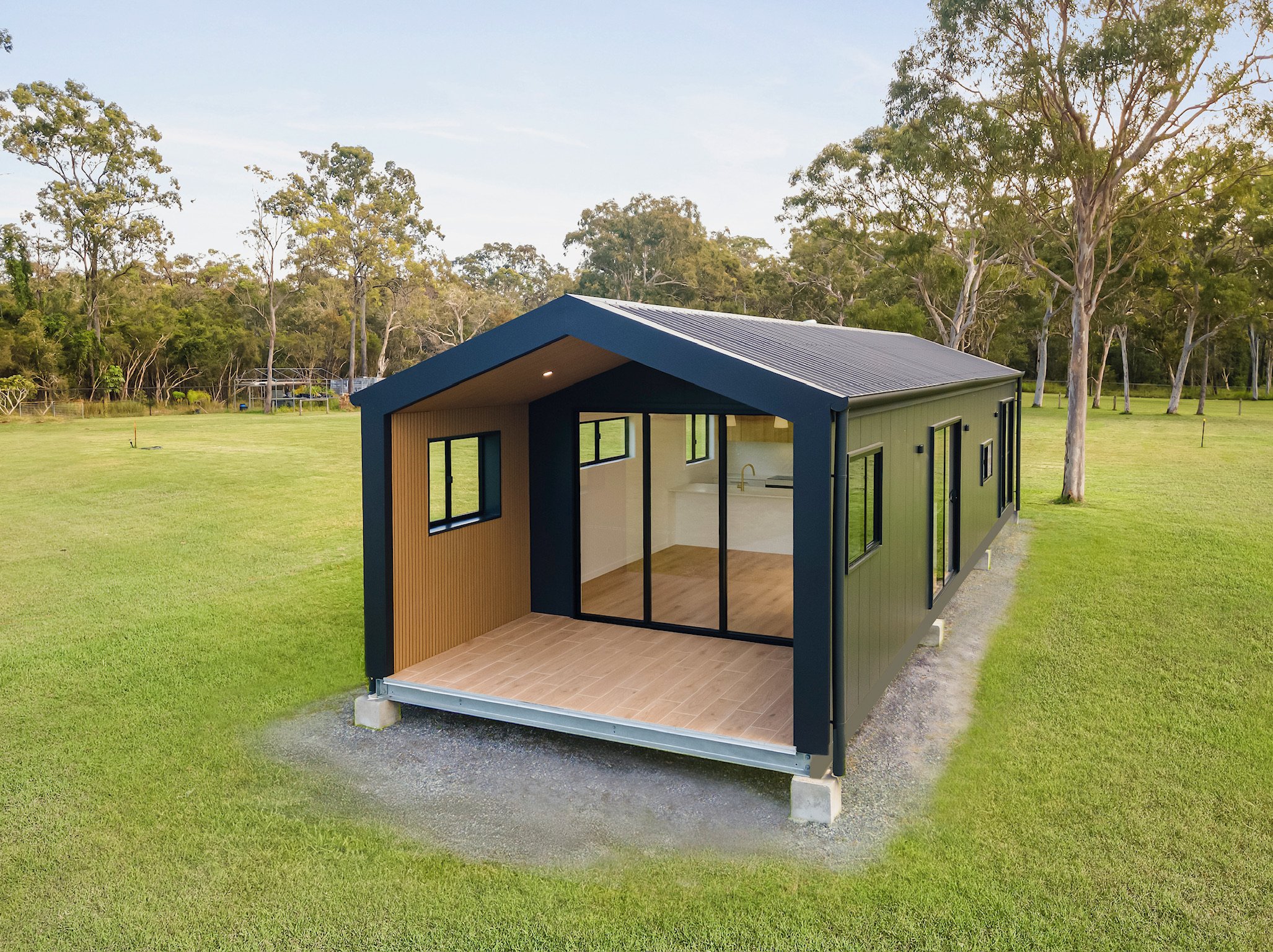 A small modern house with large glass windows and a green exterior, situated on a grassy field with trees in the background under a clear sky.
