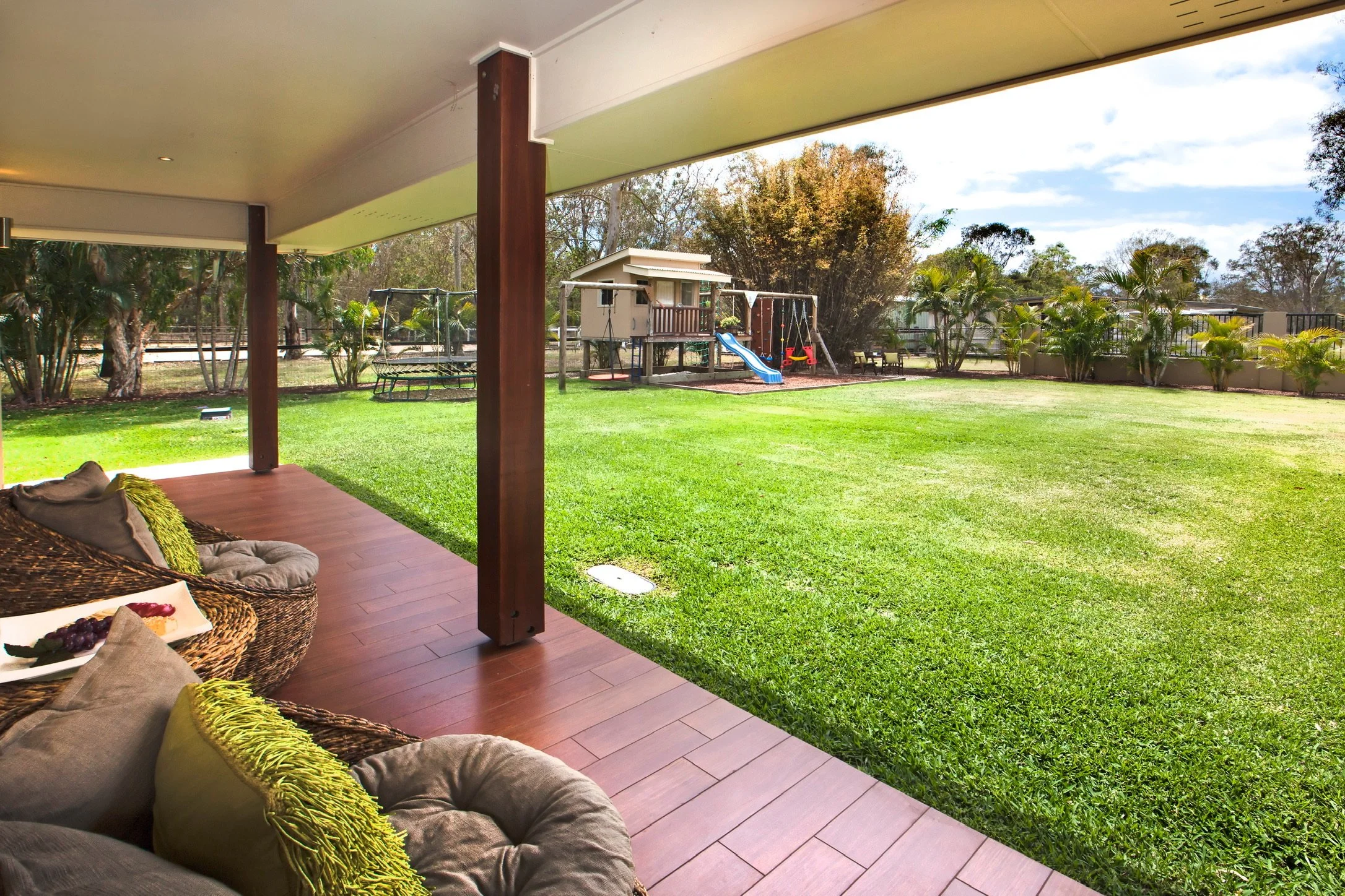 View from a covered porch overlooking a lush green backyard with a children's playset, slide, swings, and a small house with trees and a partly cloudy sky in the background.