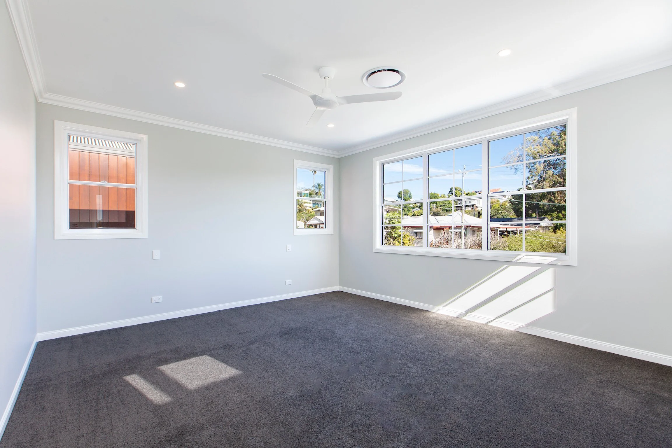 Empty room with large windows, white walls, ceiling fan, and dark carpet flooring.