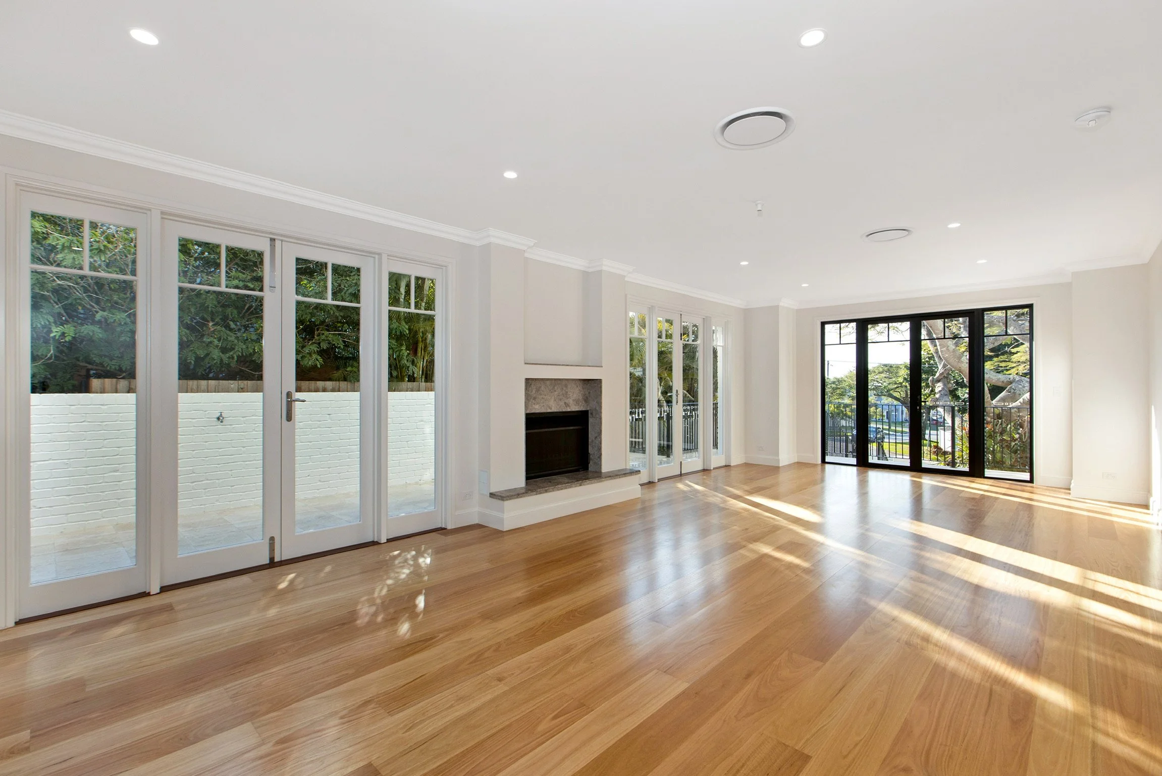 Sunlit living room with large windows and wood flooring, featuring a fireplace with a stone mantel, white walls, and black sliding glass doors leading outside.