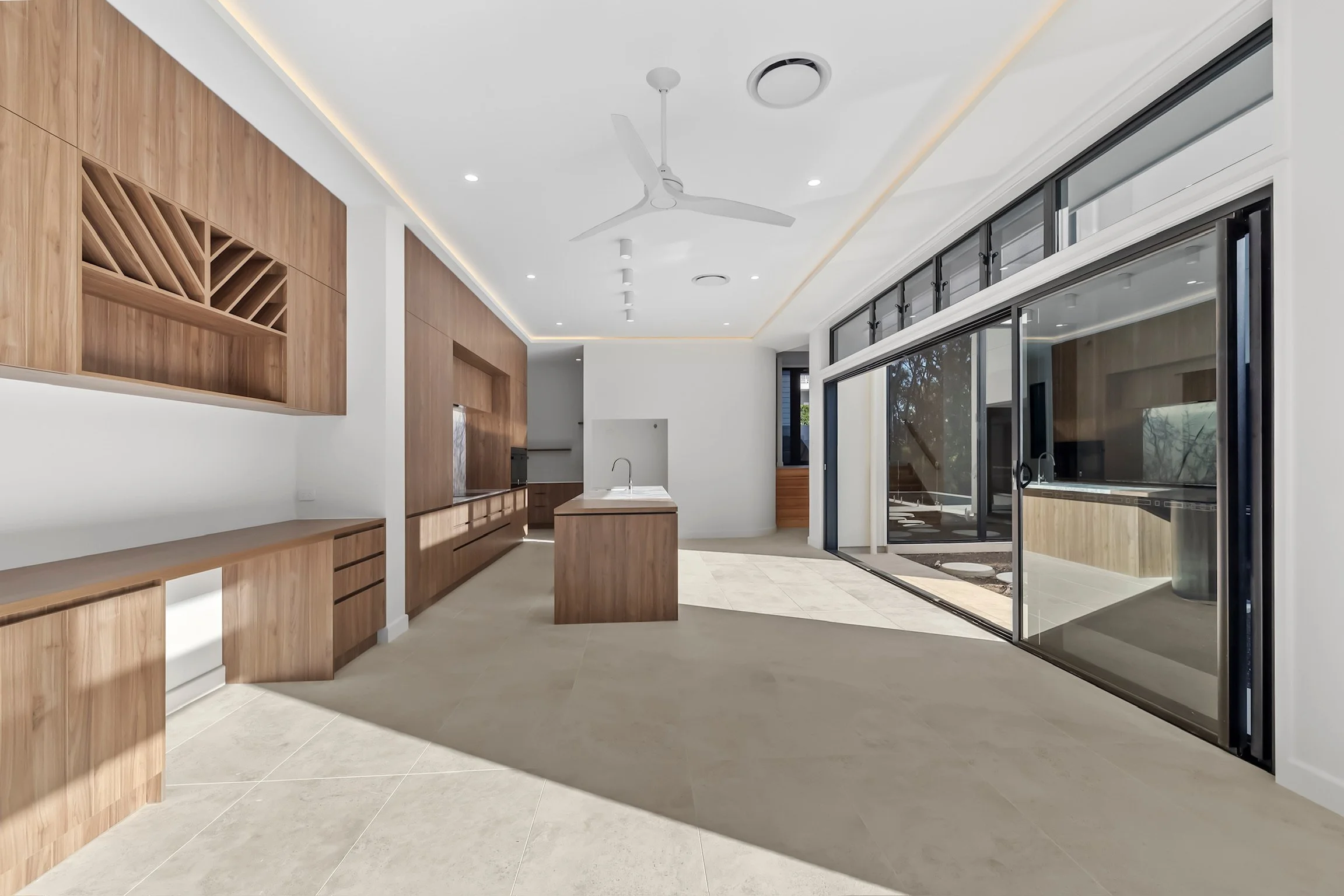 Modern kitchen with wood cabinets, a white island, and large glass sliding doors leading outside; ceiling fan and recessed lighting