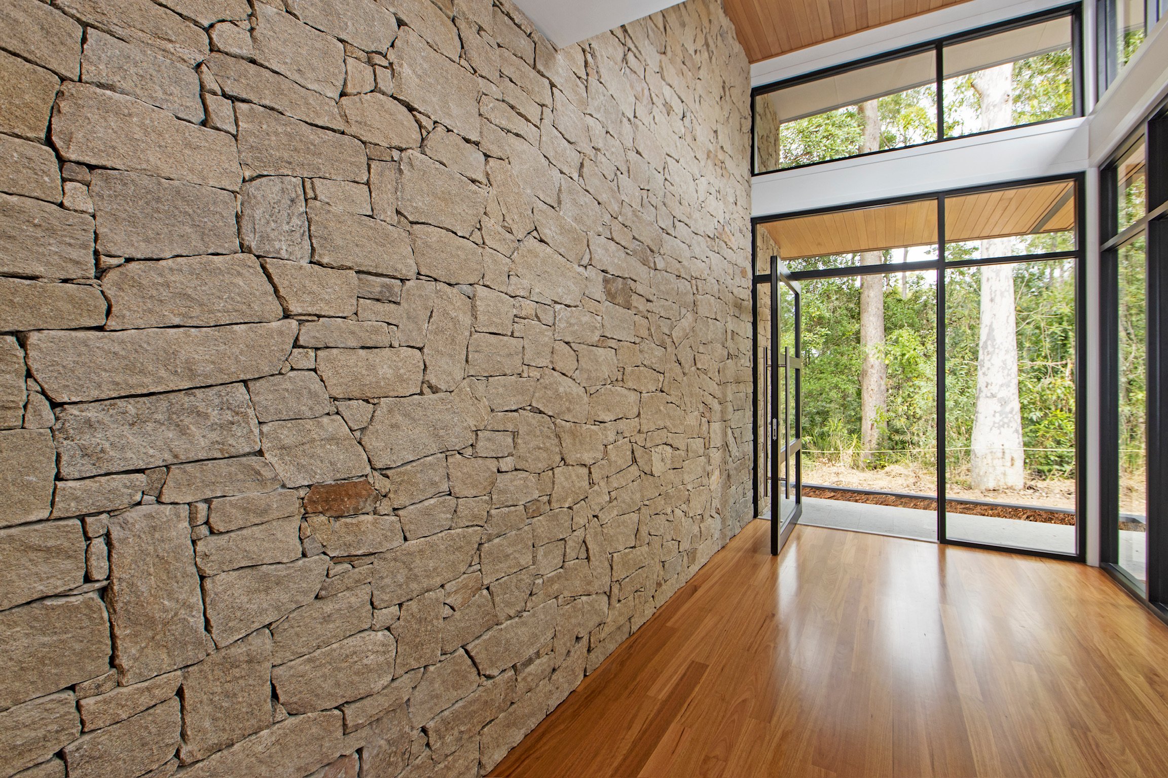 Interior view of a modern house with a stone wall, wooden flooring, and large glass windows showing trees outside.