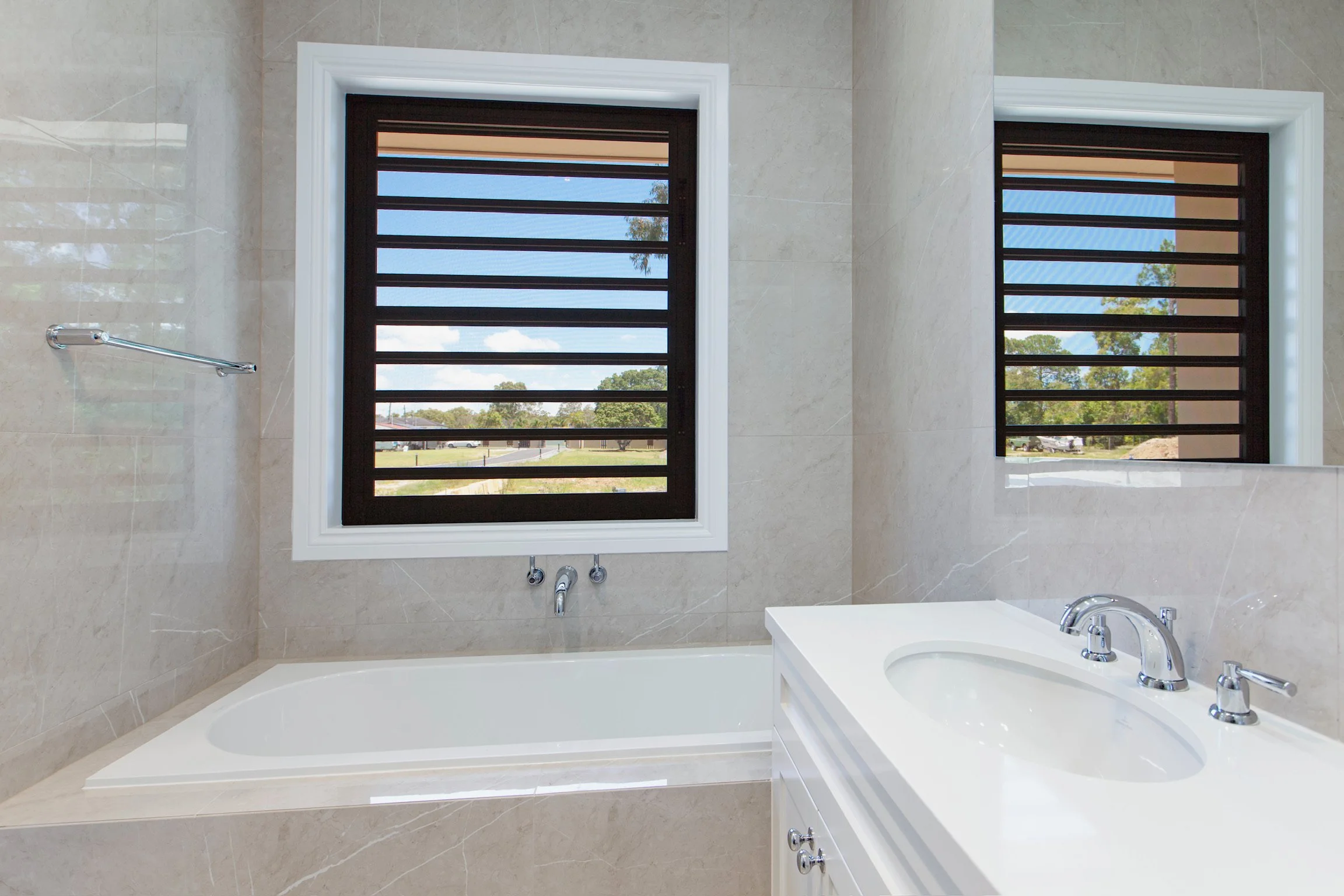 A bathroom with two window openings with black frames and horizontal bars, allowing a view of trees and blue sky outside. The bathroom has beige marble walls, a white bathtub, and a sink with a chrome faucet.