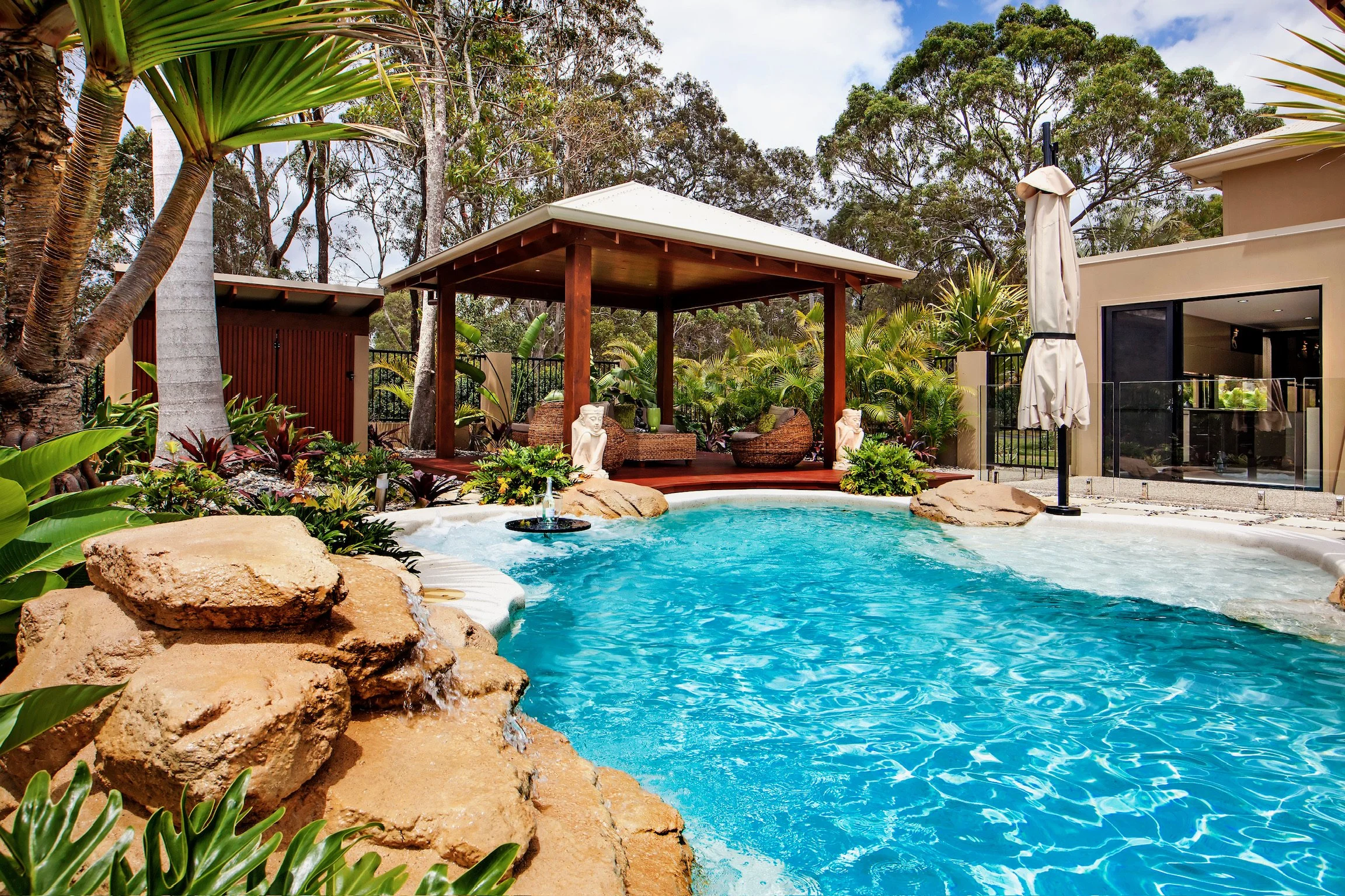 Swimming pool with rocks surrounding it, a wooden gazebo with seating, lush green plants, trees, and part of a modern house in the background.