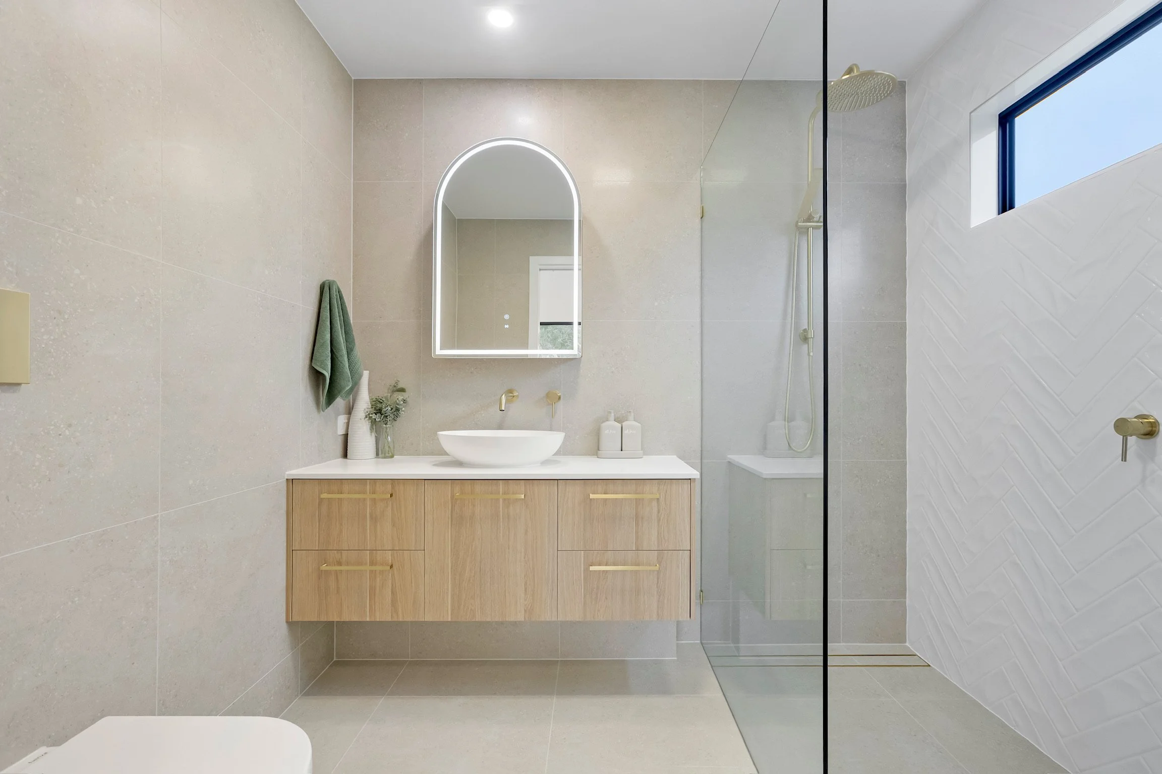 Modern bathroom with beige tiled walls and floor, illuminated mirror above a wooden vanity with a white vessel sink, green towel hanging on the wall, and a shower area with a rain showerhead and a window with a blue frame.