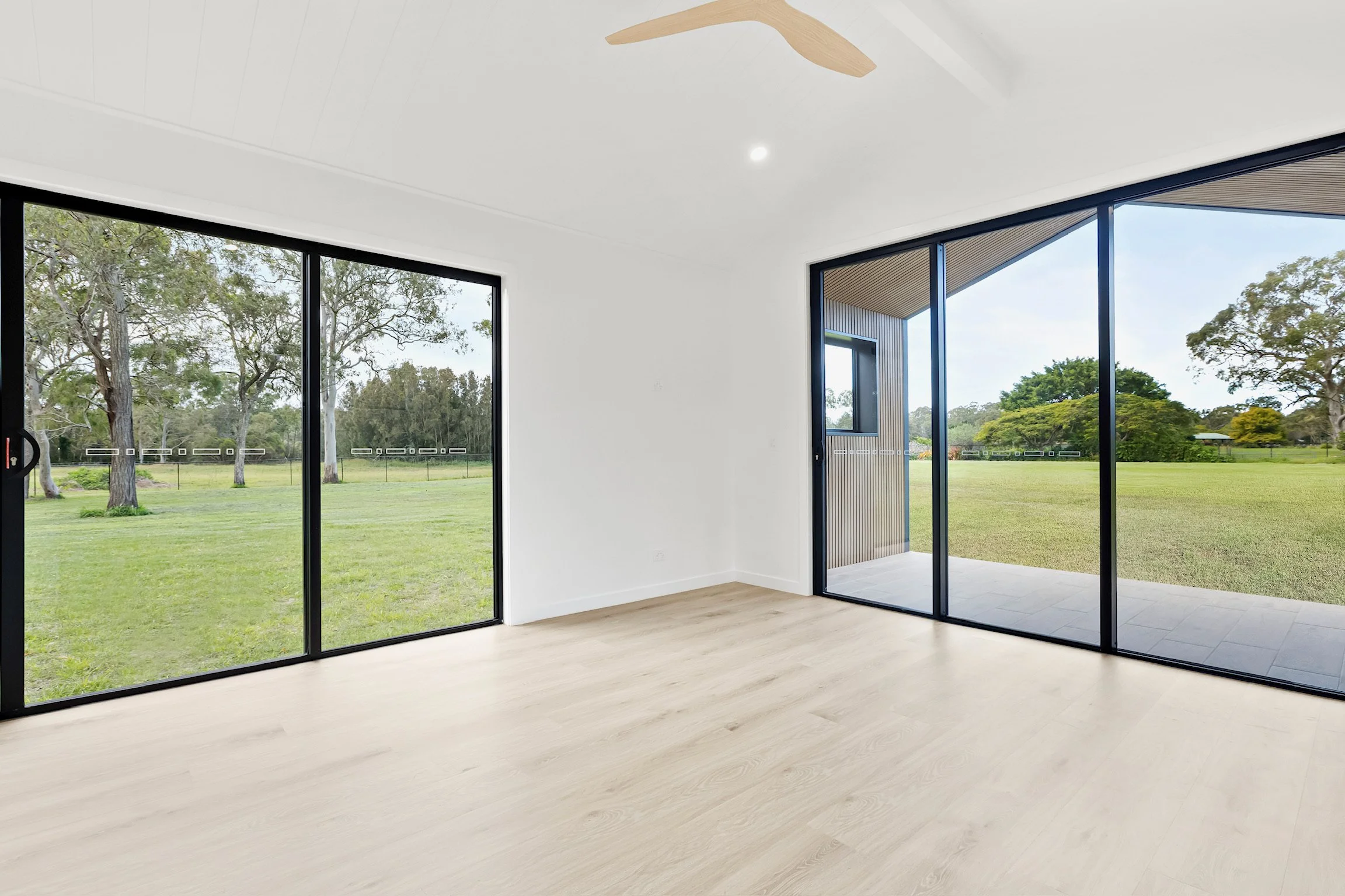 An empty room with large glass sliding doors and windows, showing a green outdoor landscape with trees and grass.
