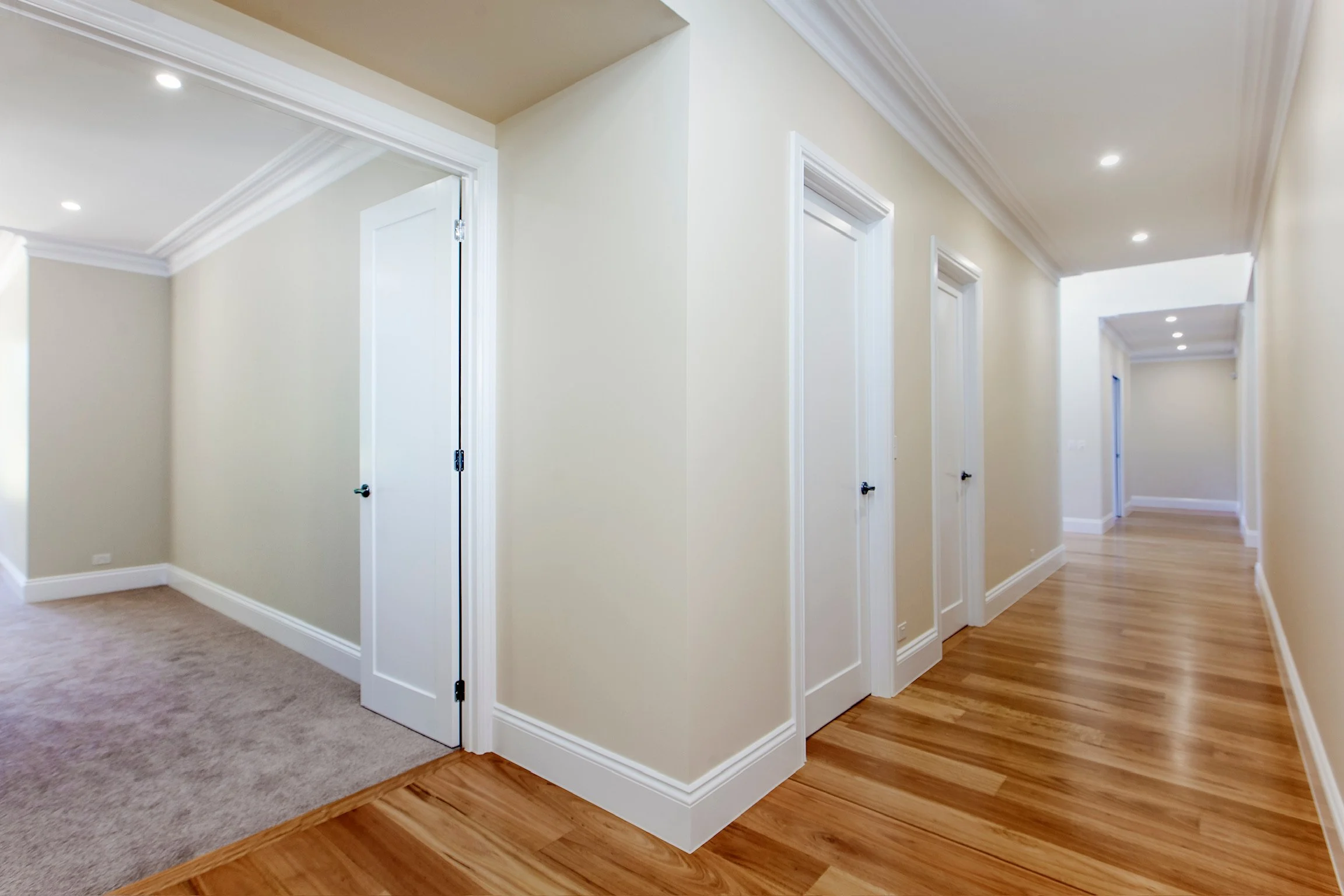 Interior view of a hallway with white walls, multiple closed white doors with black handles, crown molding, recessed lighting, hardwood flooring, and a section of beige carpet.