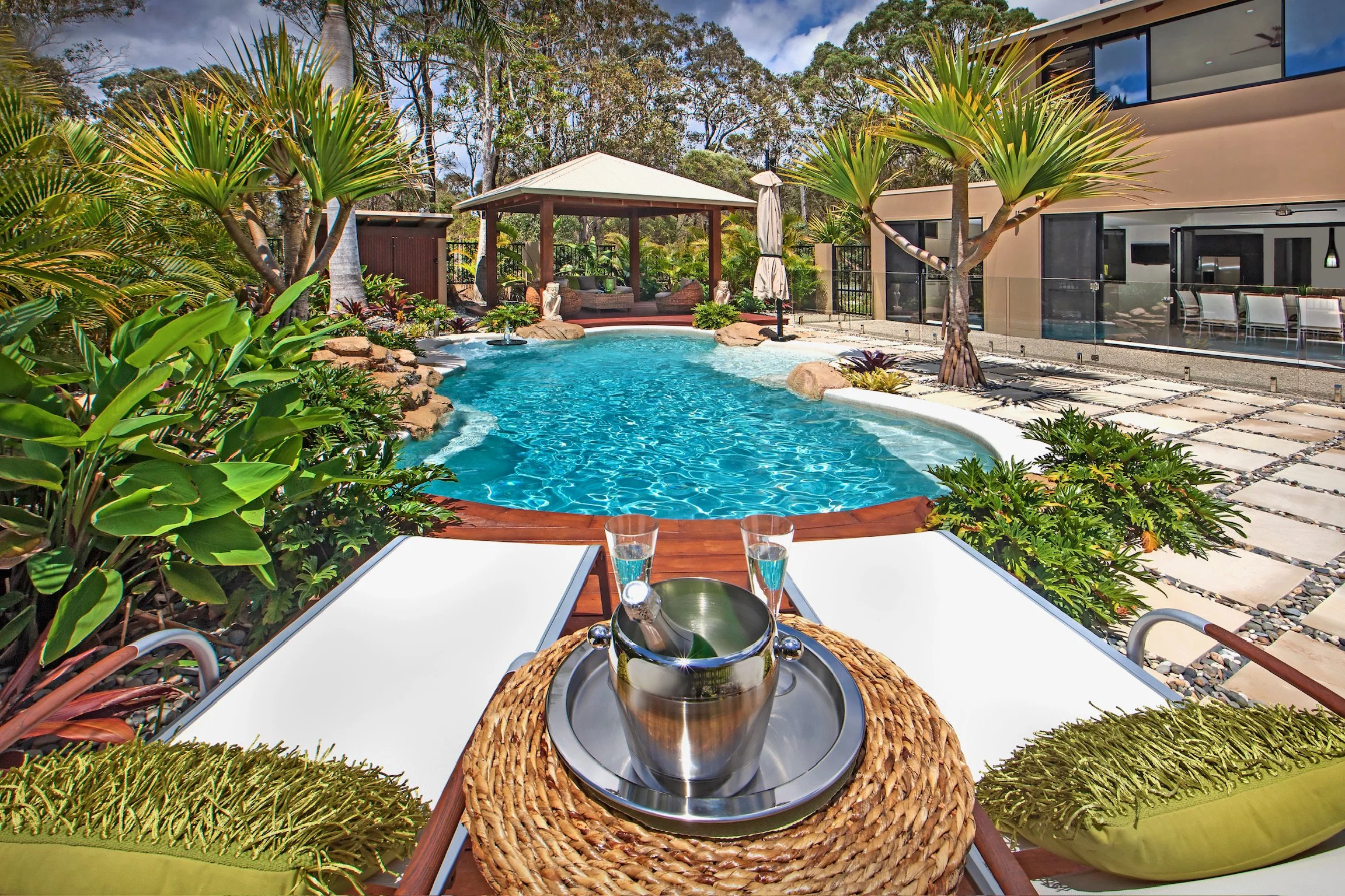 A backyard with a swimming pool surrounded by tropical plants, trees, and outdoor furniture. A small table with a wine bucket and glasses is in the foreground.