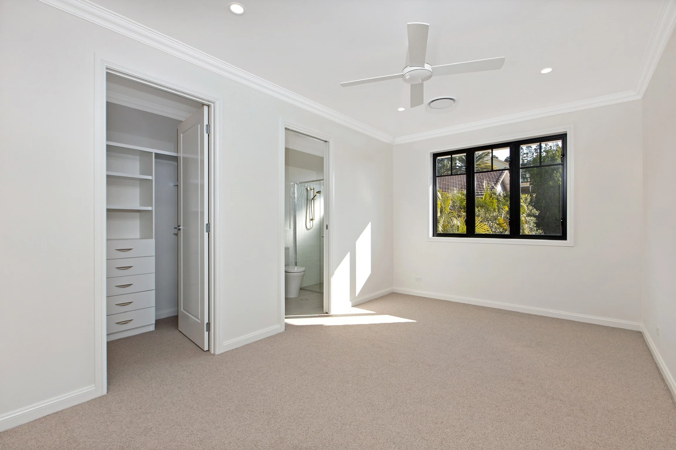 Empty bedroom with cream-colored carpet, white walls, a large window, and an open closet with shelves and drawers. A view of neighboring houses and trees through the window.