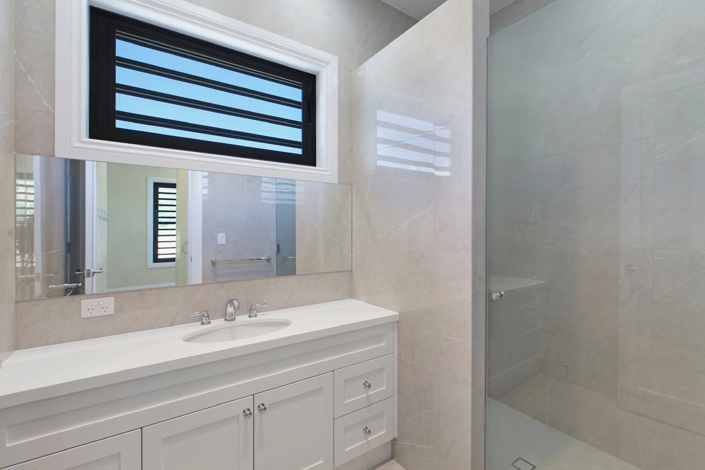 Bathroom with a large mirror, white vanity with multiple drawers, two faucets, a tiled wall, a window with black blinds, and a glass shower door.
