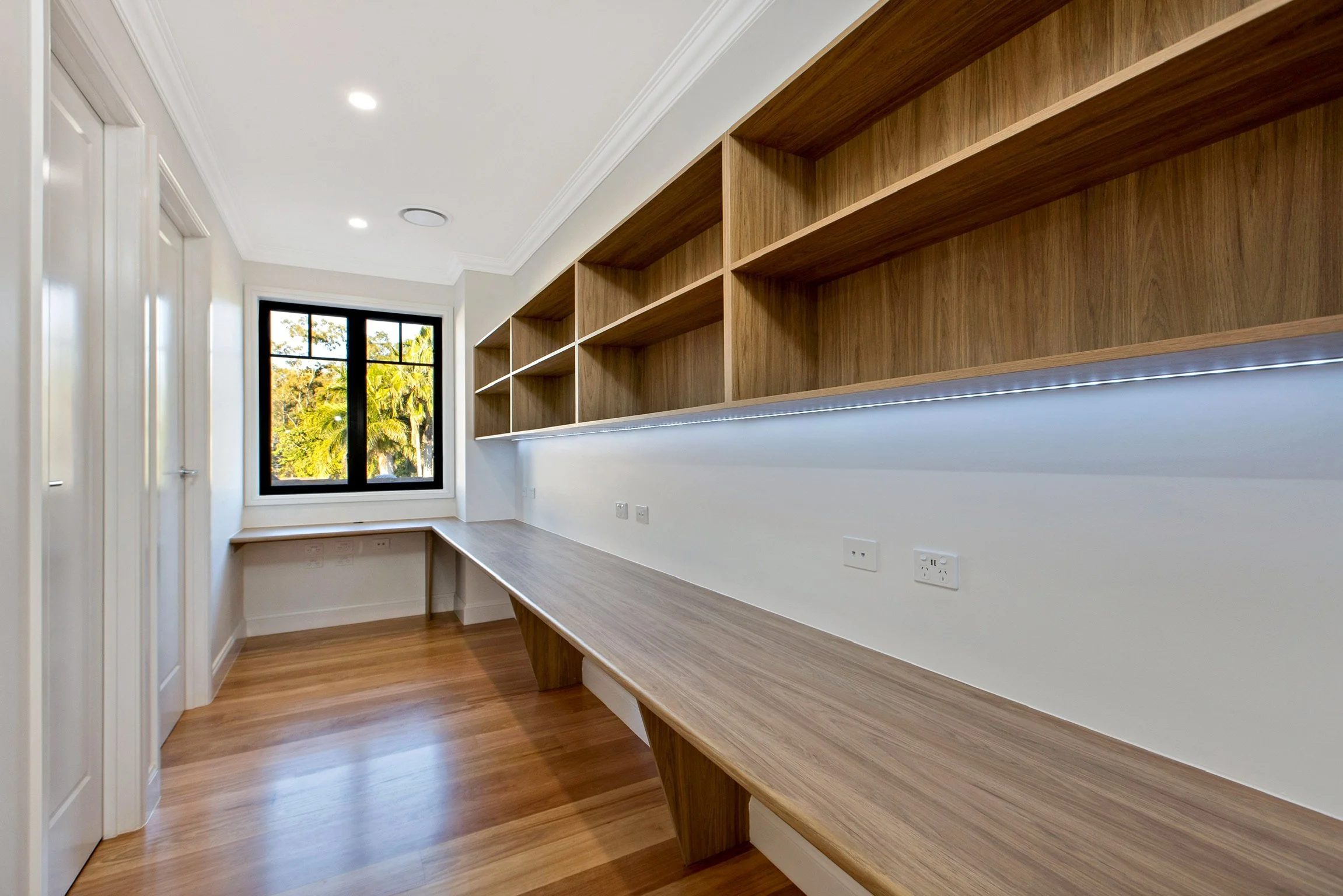 Room with built-in wooden shelving and a desk, featuring a window with a view of trees outside, white walls, and recessed lighting.