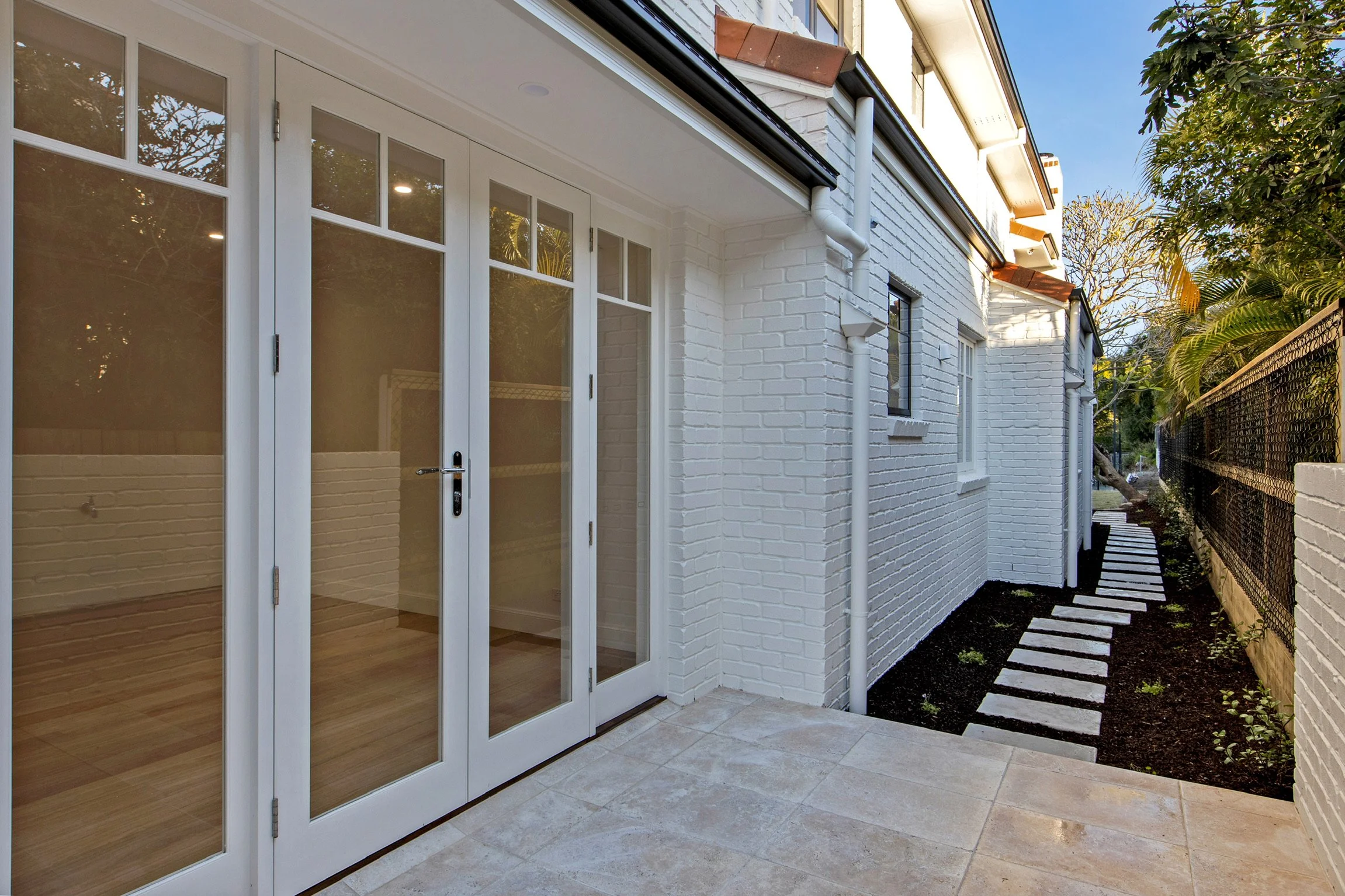 Backyard patio area with white brick exterior walls, glass double doors, tiled flooring, a pathway made of stepping stone slabs, and lush green trees and plants along the fence.