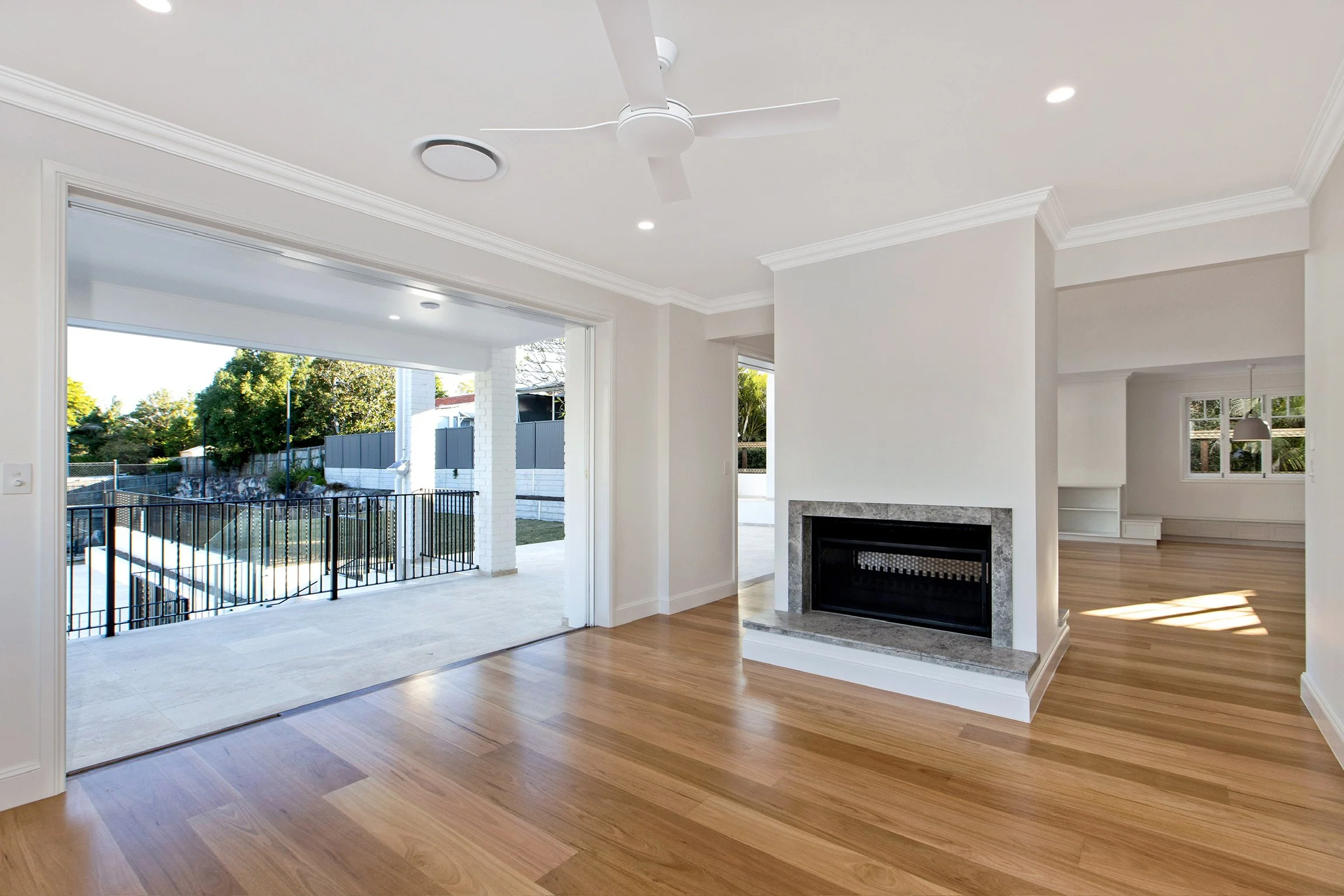 Interior of a modern house with hardwood floors, white walls, a fireplace with a stone surround, a ceiling fan, and an open sliding door leading to a backyard with a fence and trees.