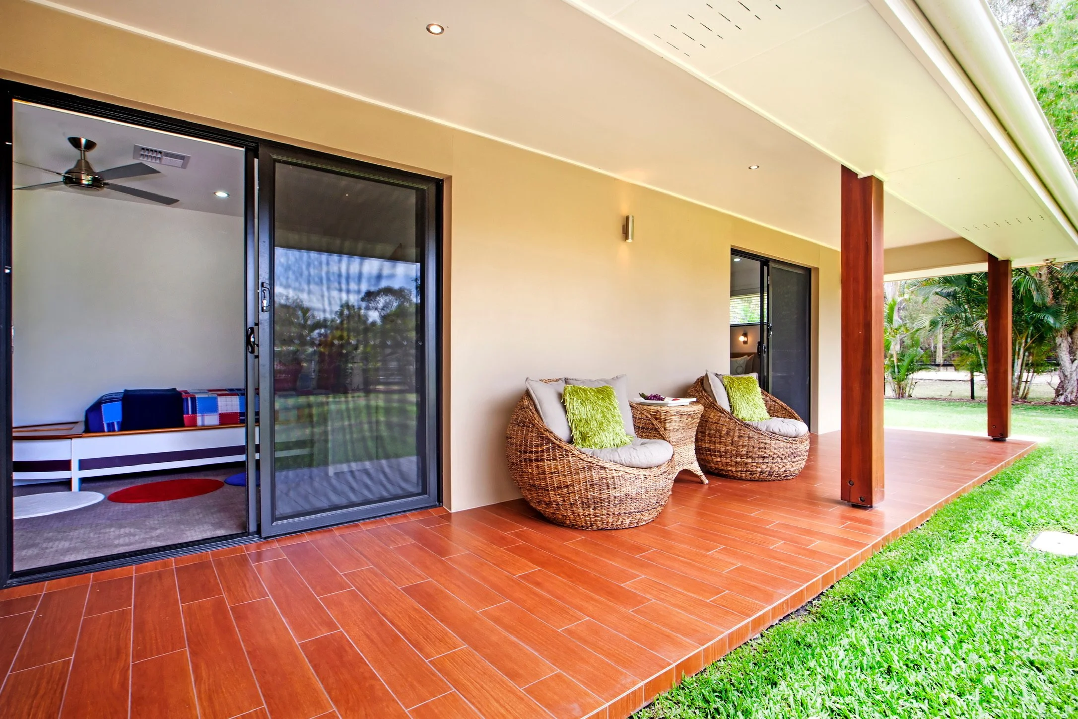 A covered outdoor patio area with two wicker chairs with white cushions and green throw pillows. There is a small table with decorative items between the chairs. The patio has a hardwood floor and is next to a grassy yard with trees. Two sliding glas