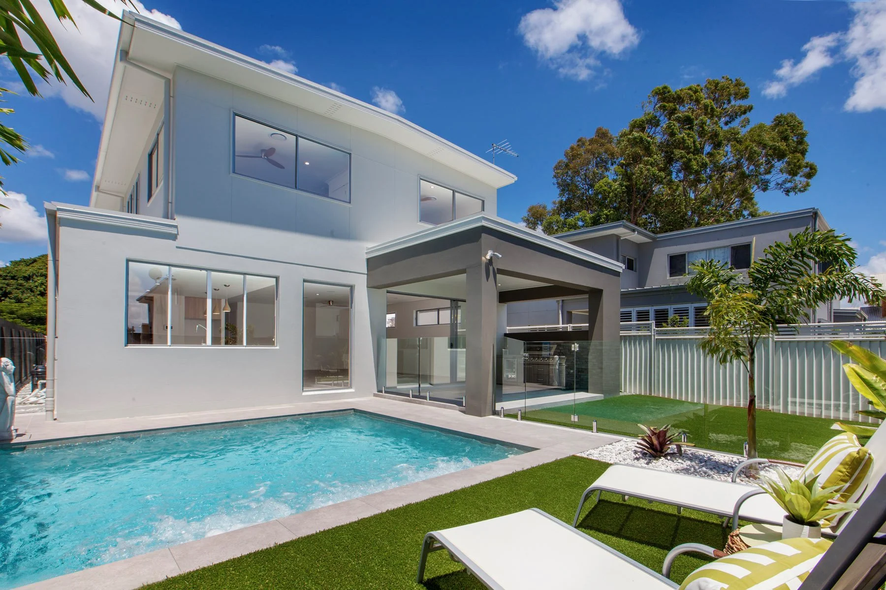 Modern two-story house with large windows, a swimming pool, and a backyard with lounge chairs and plants, under a blue sky with some clouds.