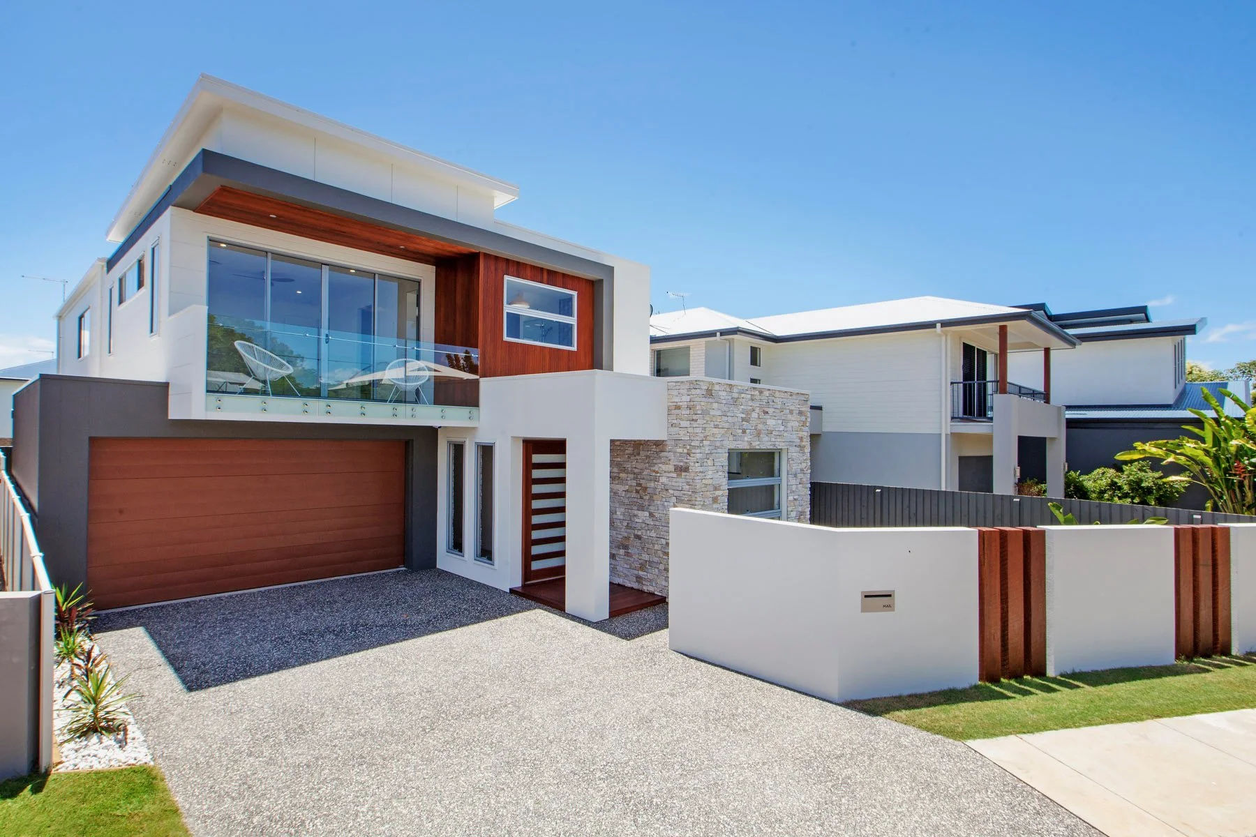 Modern two-story house with a wooden garage door, large glass balcony, stone accent wall, and white exterior walls, under a clear blue sky.