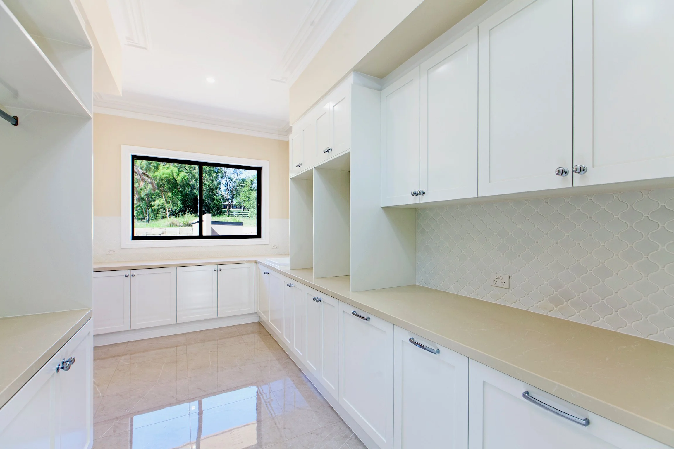 Empty laundry room with white cabinets, a window showing greenery outside, and beige tiled flooring.