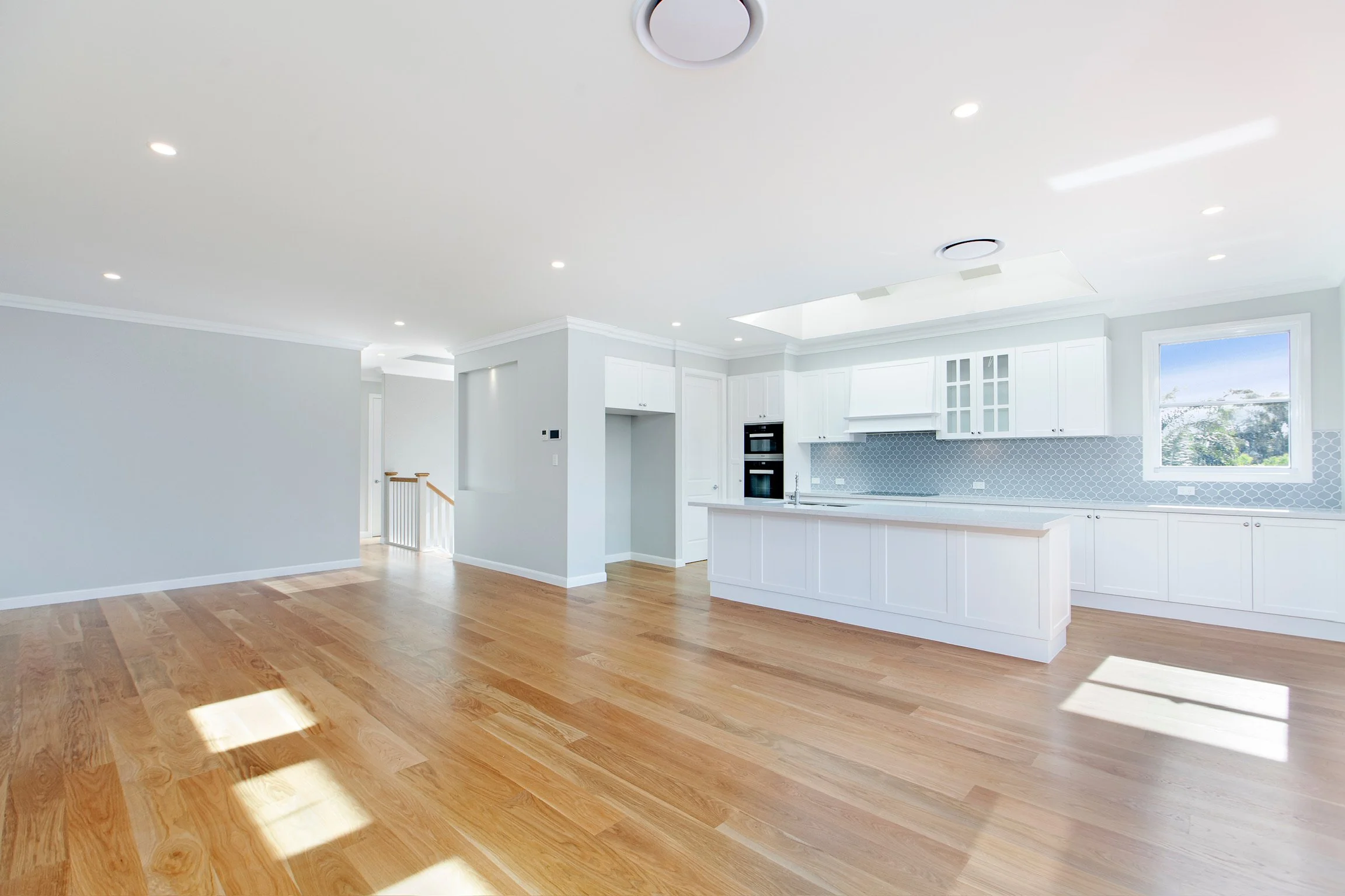 A bright, modern kitchen with white cabinets, a blue hexagonal tile backsplash, large windows, and wood flooring.