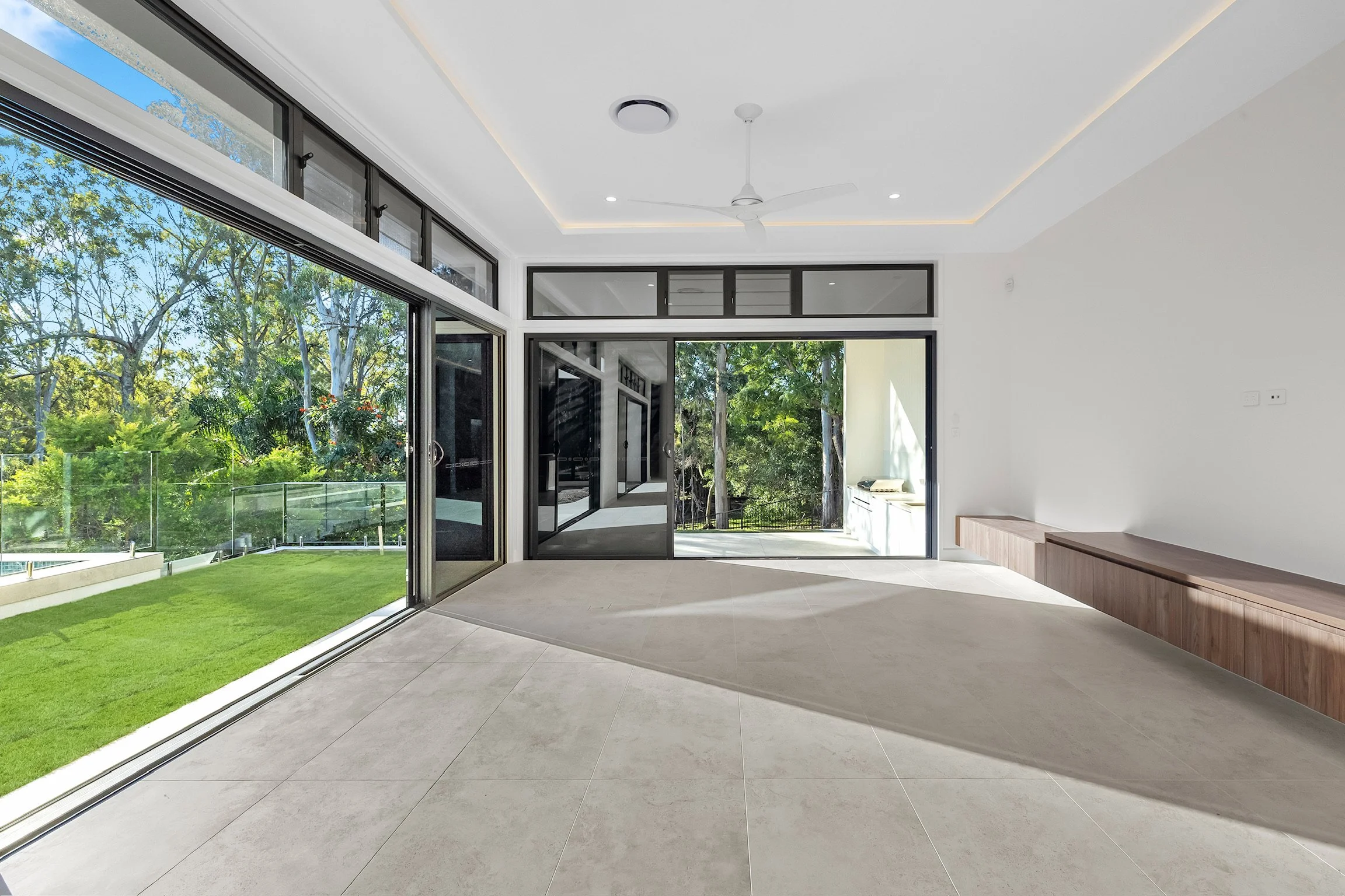Modern interior room with large glass sliding doors opening to a green backyard with trees. The ceiling is white with recessed lighting and a ceiling fan. The floor is tiled in light gray, and there is a built-in wooden bench on the right side.
