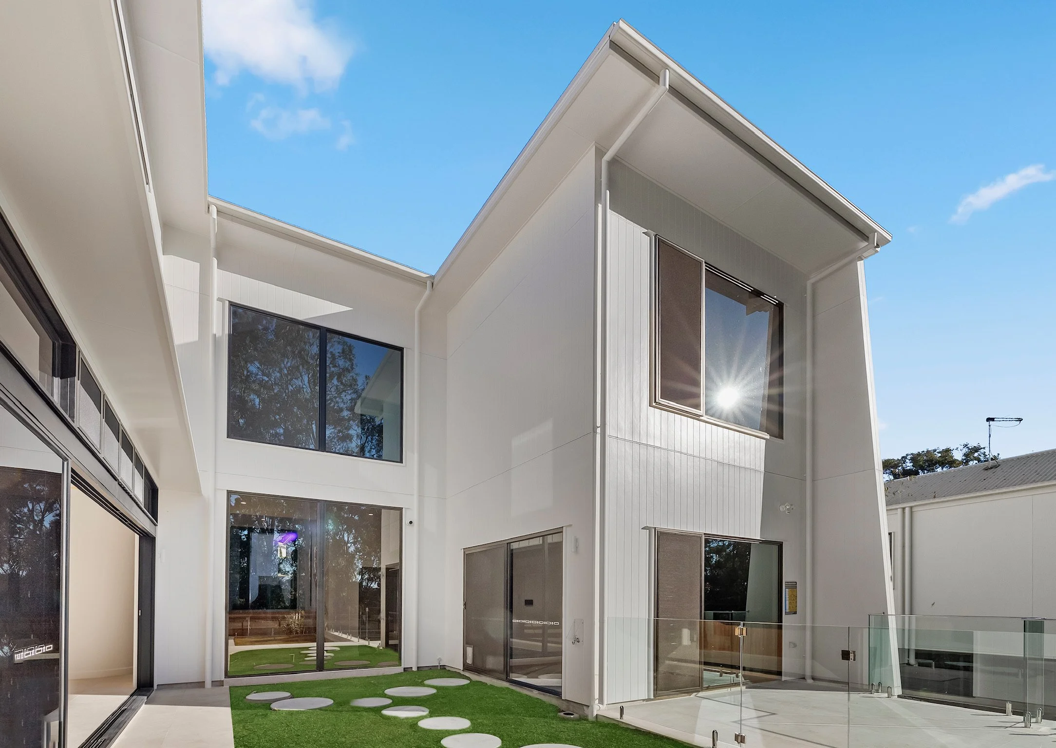 Modern two-story white house with large windows, a small grass courtyard with stepping stones, and a clear blue sky overhead.