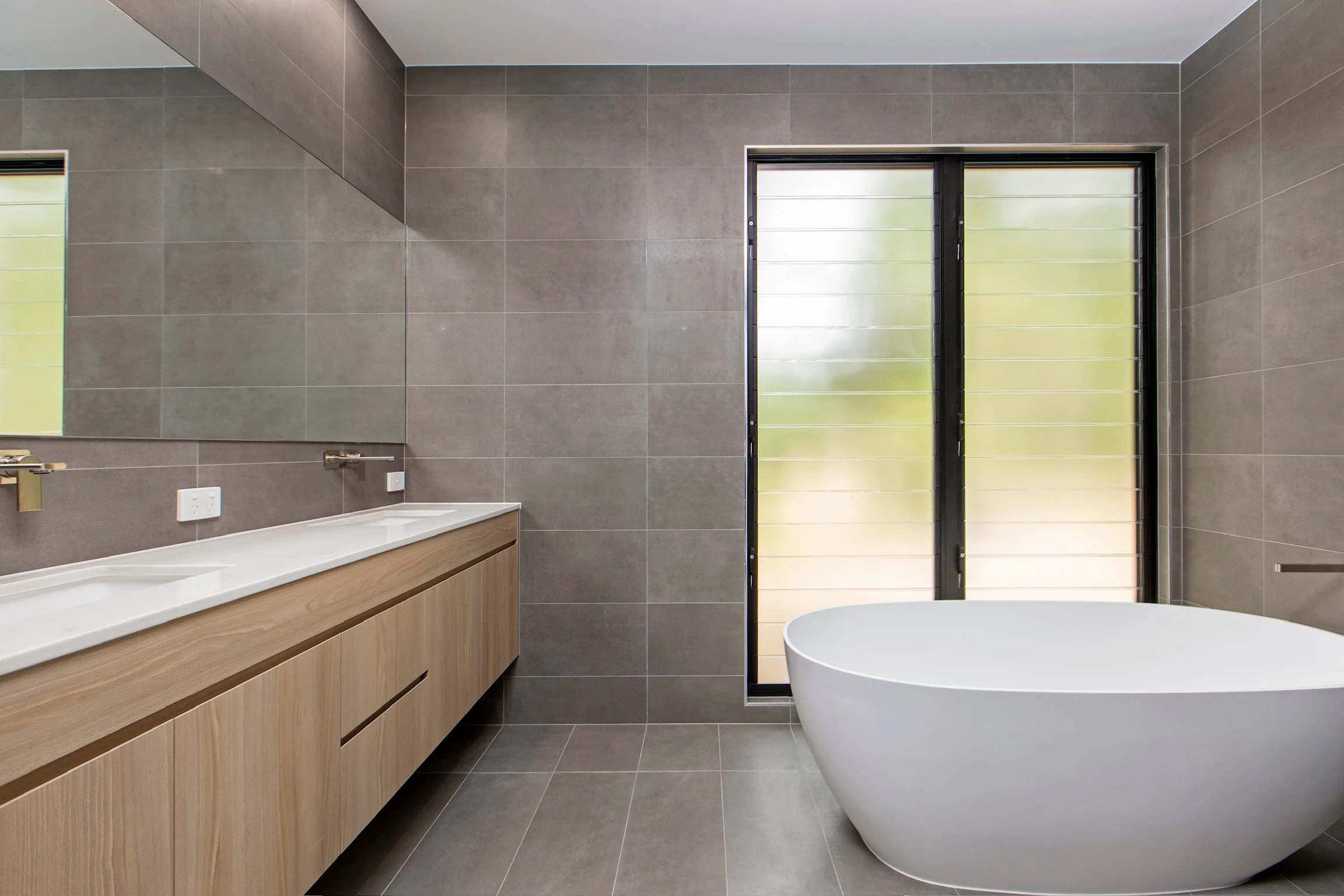 Modern bathroom with gray tiled walls and floor, a wooden vanity with a white countertop, a mirror, a window with black framing, and a white freestanding bathtub.