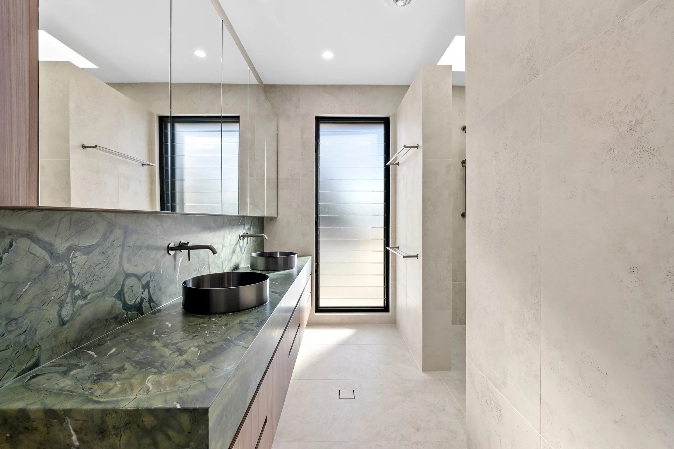 Modern bathroom with beige walls, a large mirror, two black vessel sinks, and a long green marble countertop. There is a window with black frames and frosted glass, and a door with the same black framing. The floor and walls are tiled in neutral tone