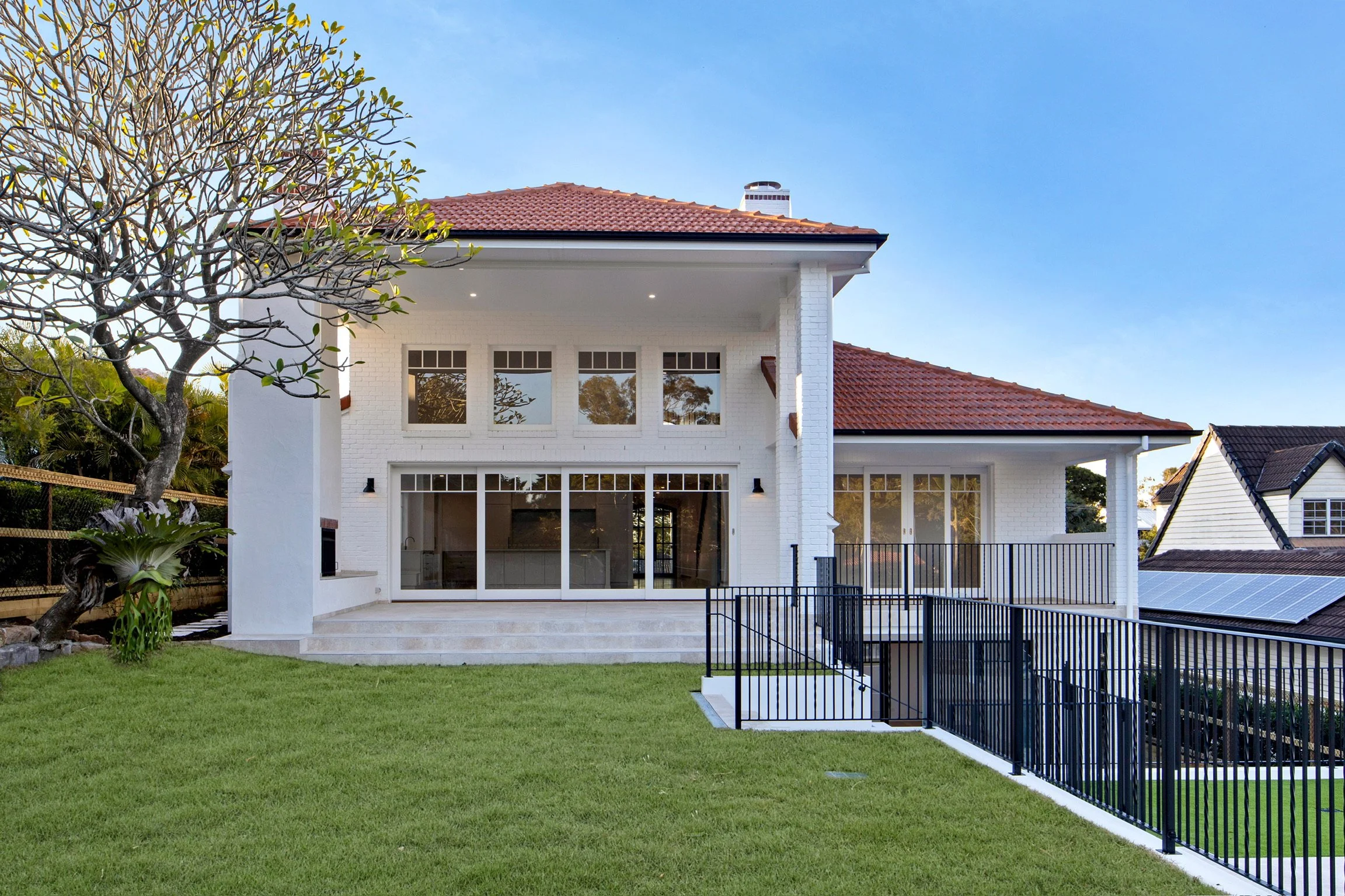 Front view of a modern two-story house with white brick walls and a red tiled roof, large windows, a green backyard with grass, a tree on the left, and a black metal fence in the foreground.