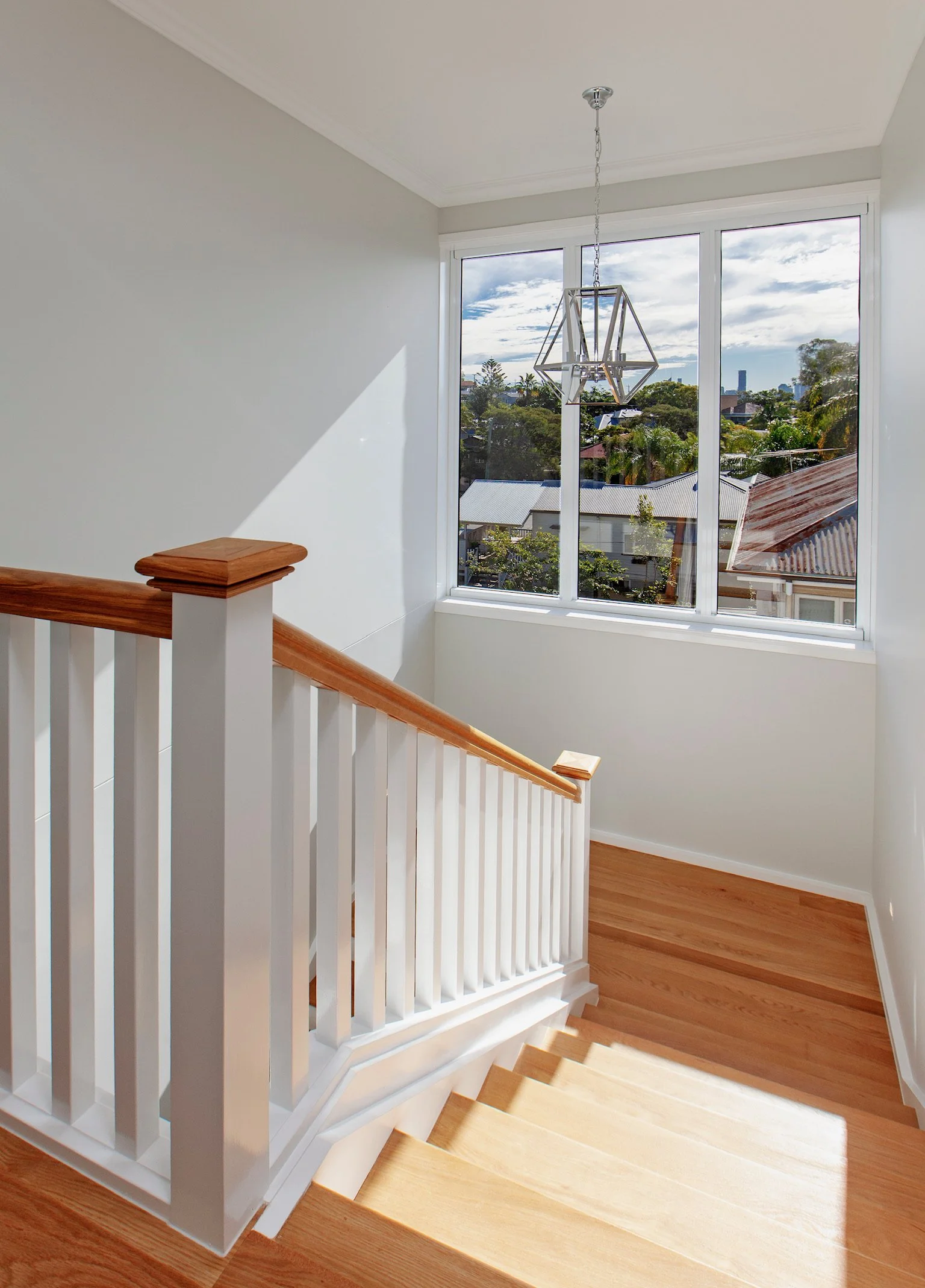 Interior view of a staircase with a white railing and wooden handrail, leading down to a wooden floor, overlooking a bright room with large window showing a view of trees, rooftops, and a cloudy sky outside.