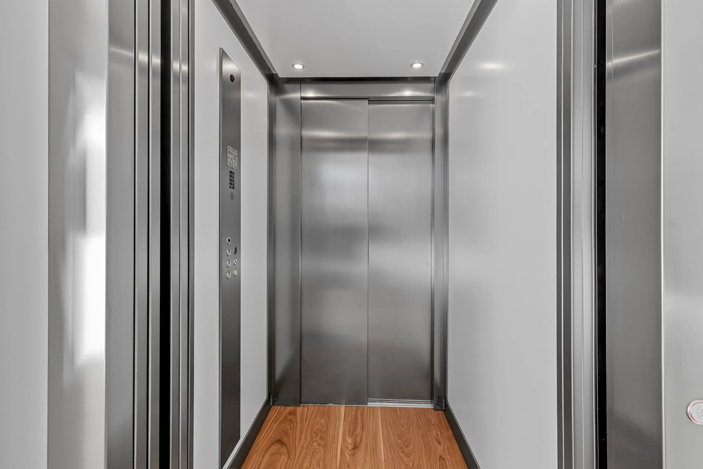 Modern elevator with closed metallic doors, elevator control panel on the left, wood flooring in front, and ceiling lights.