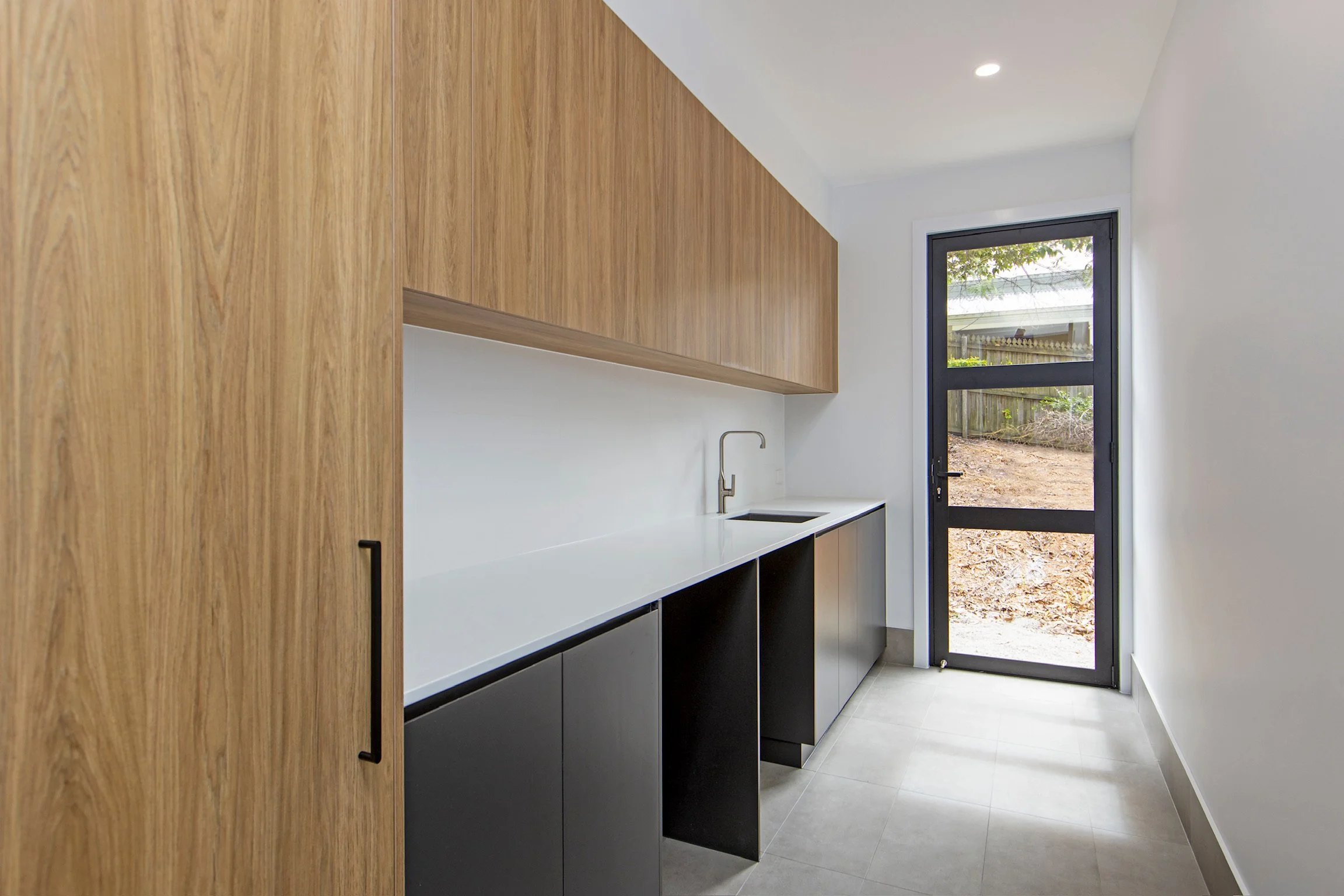 Interior view of a modern kitchen with wooden cabinets, white countertop, black lower cabinets, and a large window or door leading outside.