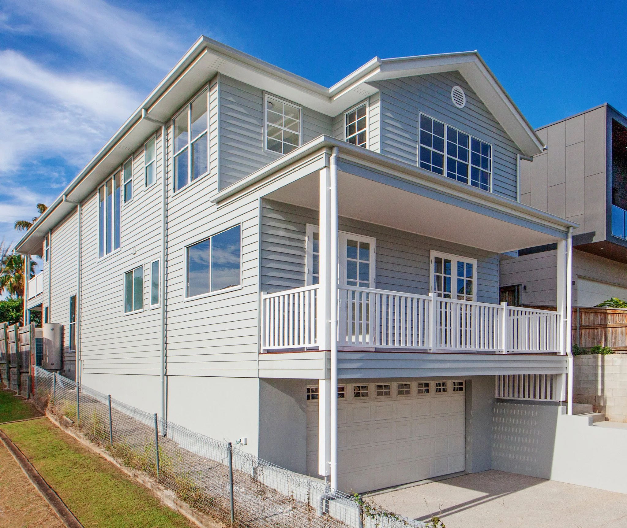 A modern, white three-story house with multiple large windows and a balcony, featuring horizontal siding and a garage door on the ground level. The house has a sloped roof and a small staircase leading to the front door. A wooden fence surrounds the yard, and there's a blue sky with some clouds overhead.