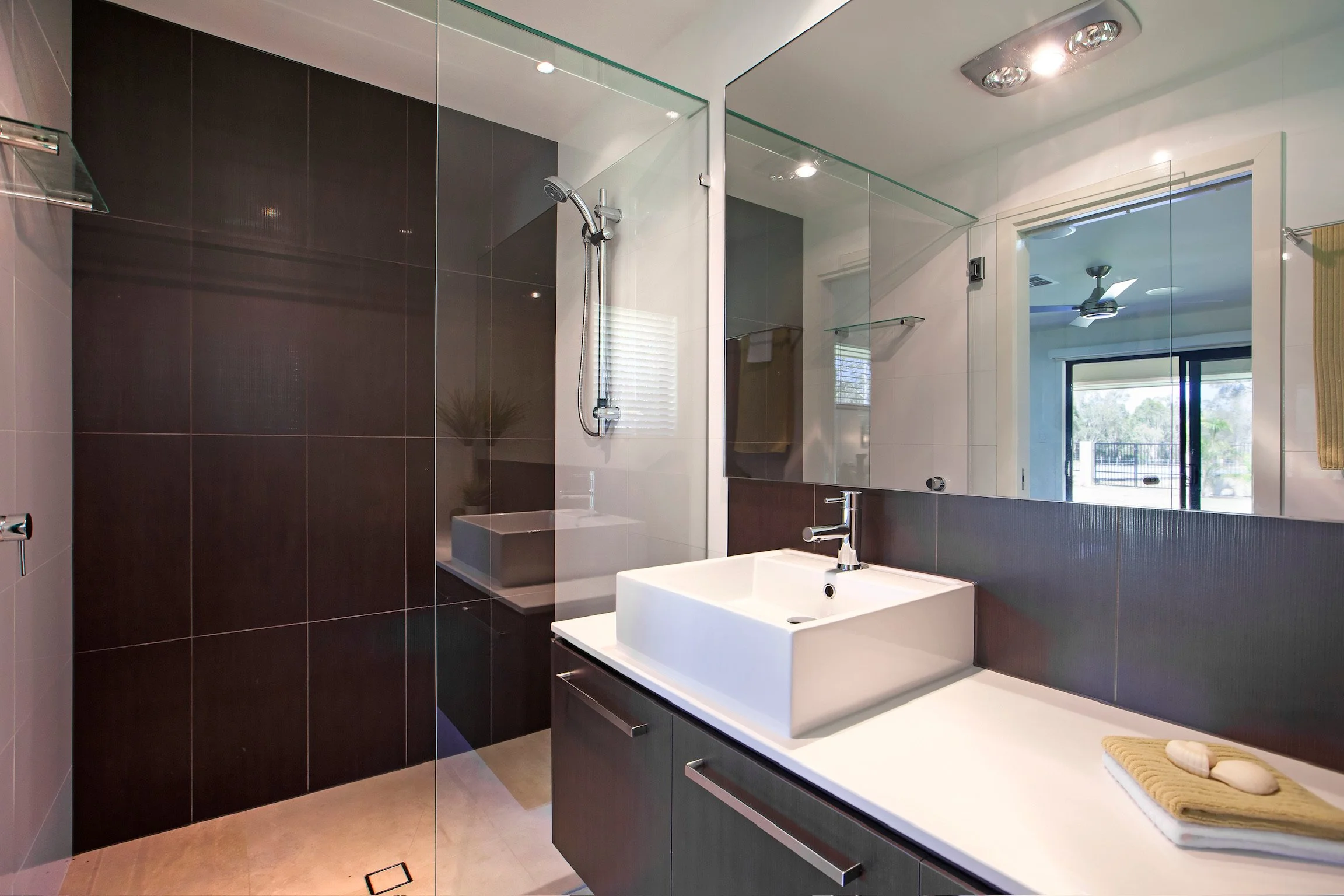 Modern bathroom with dark tiles, a white rectangular sink with a silver faucet, a large mirror, and a glass-enclosed shower area with a mounted showerhead.