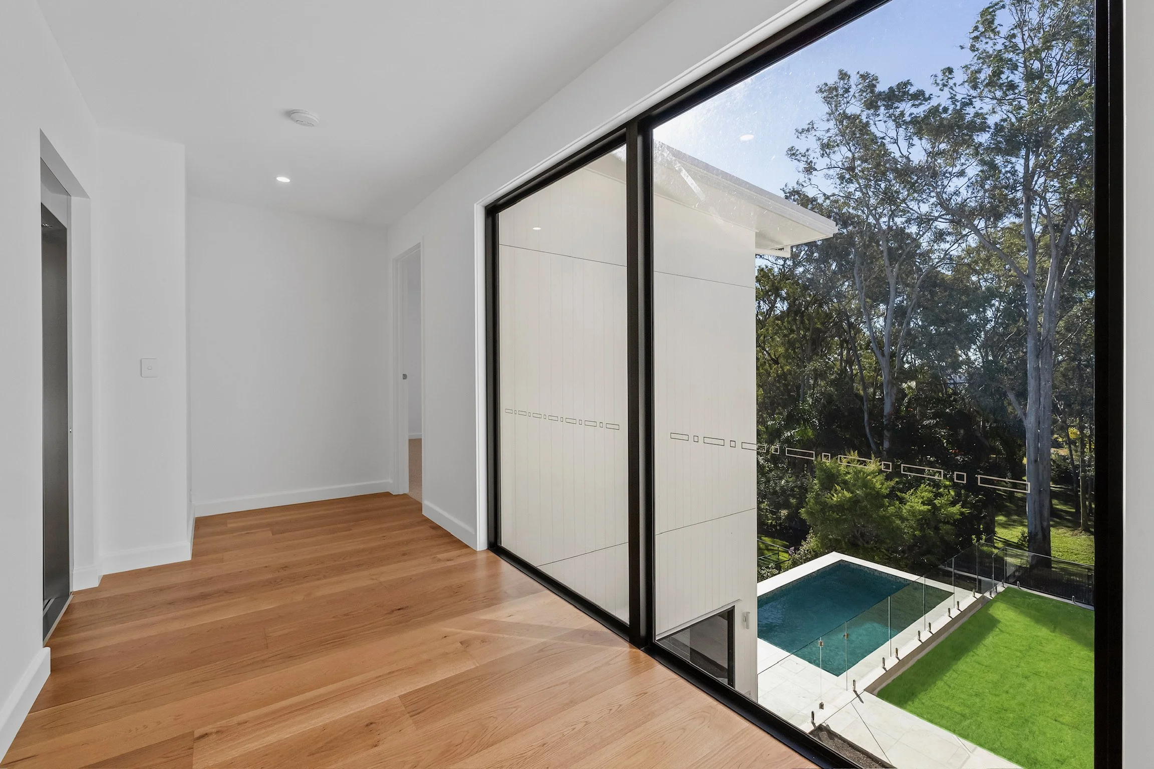 Interior view of a modern room with wood flooring, large sliding glass doors, and a balcony overlooking a backyard with trees and a swimming pool.