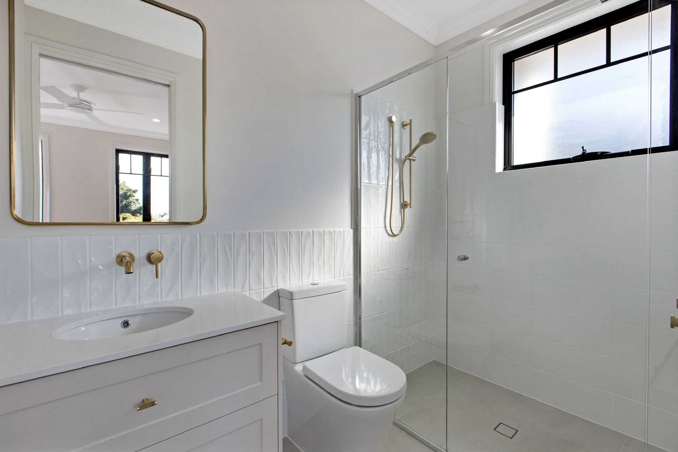 Modern bathroom with white vanity, gold fixtures, toilet, shower with glass partition, and frosted window.