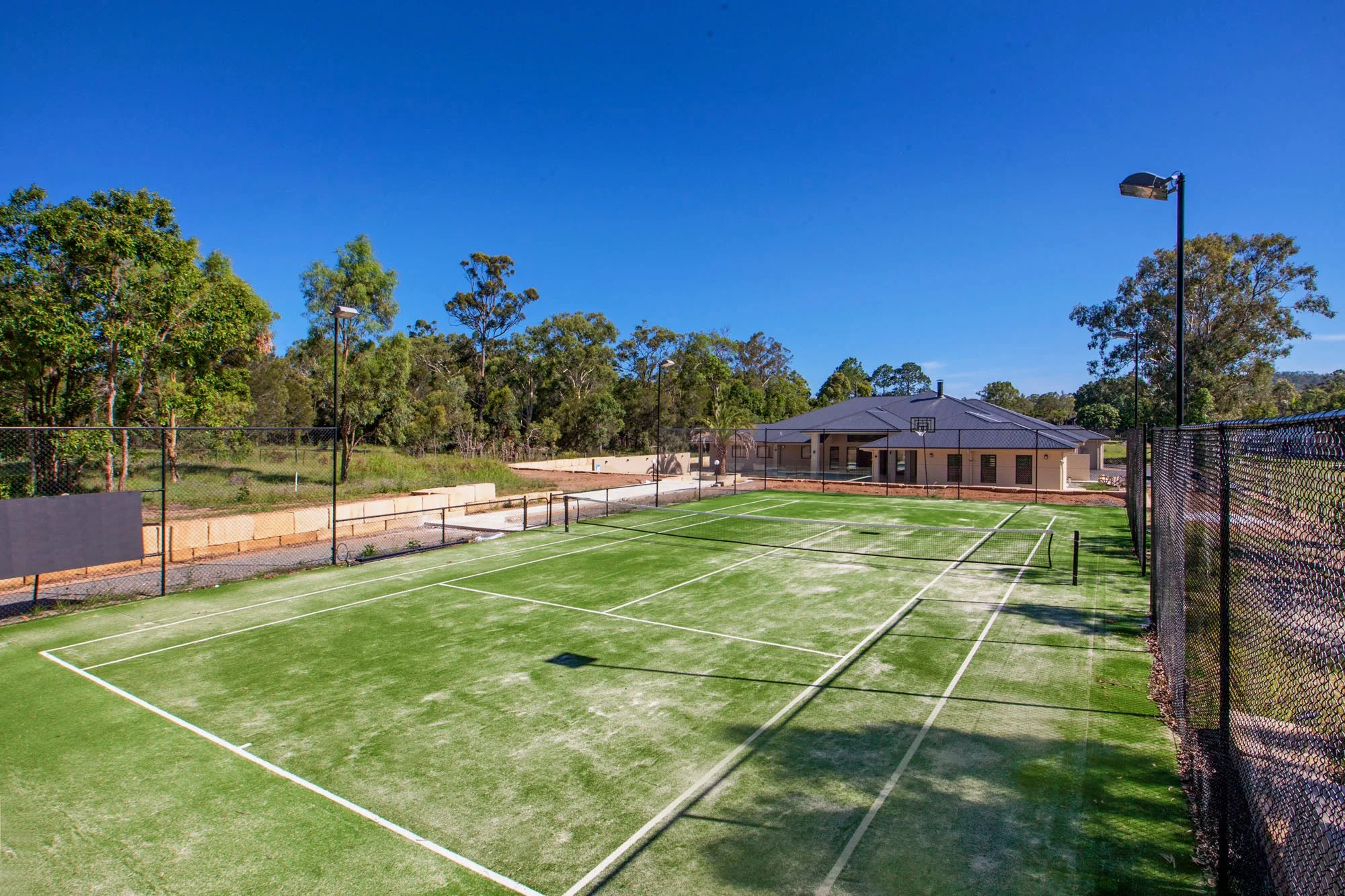 An outdoor tennis court with green surface and white lines, surrounded by a chain-link fence, with trees and a building in the background under a clear blue sky.