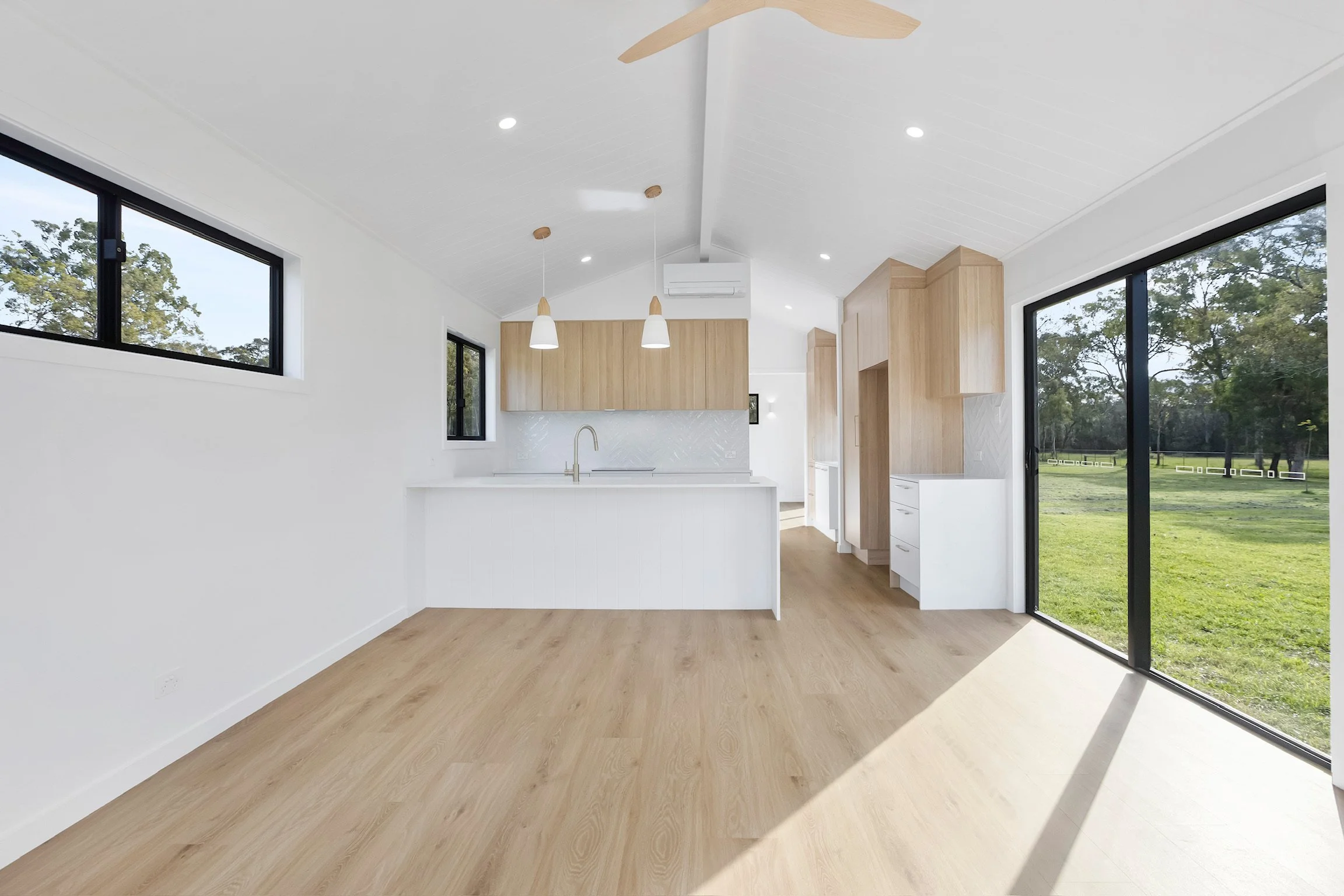 Modern kitchen with white walls, light wood cabinetry, large windows, and sliding glass doors revealing a green outdoor space.