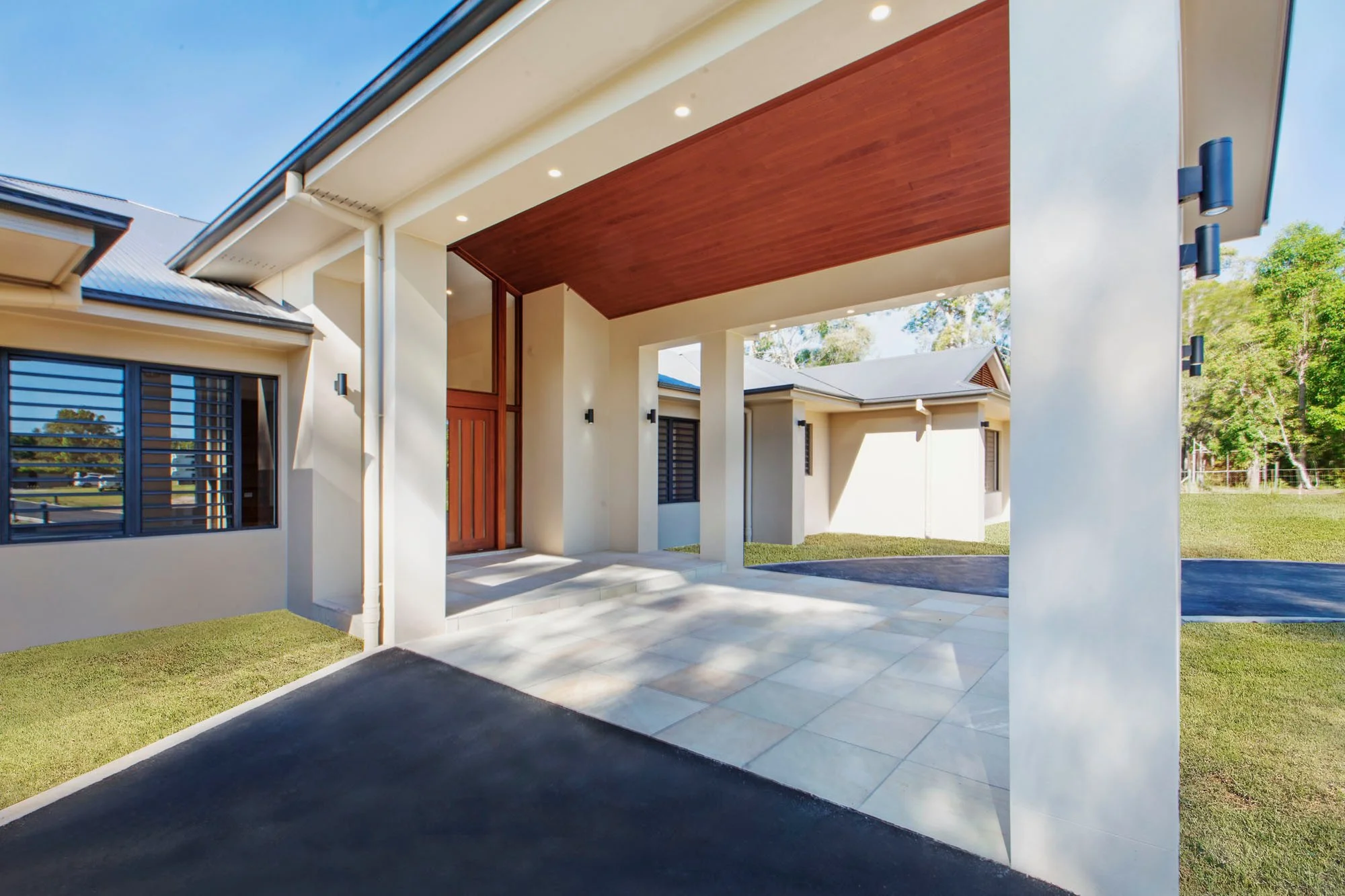 Modern house with a covered entryway, cream exterior walls, large windows, and a wooden ceiling for the porch, surrounded by a lawn and trees.
