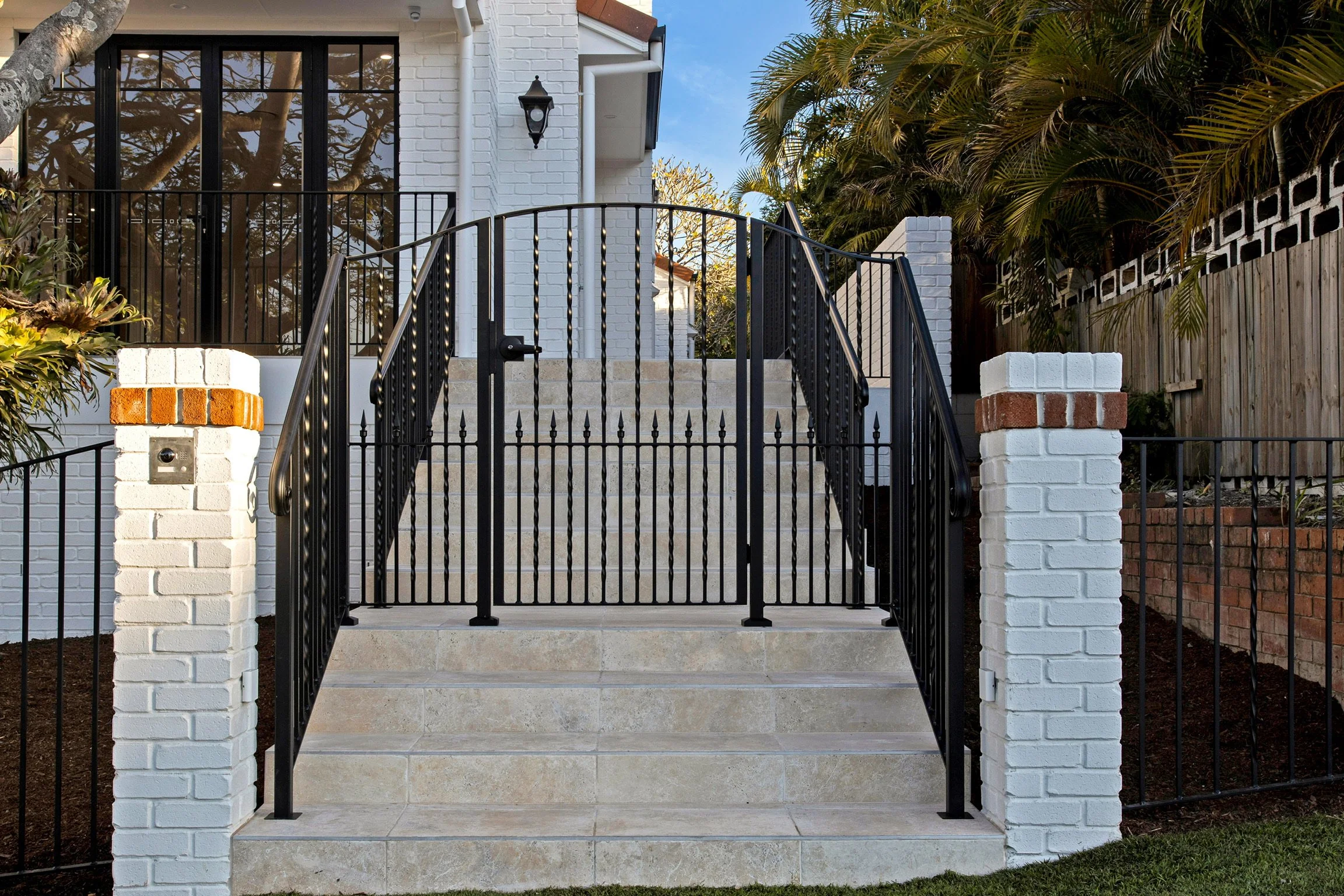 A front porch with wide beige tiled steps, black metal railing, white brick columns, and a white brick house with a black window and a black outdoor lantern, surrounded by greenery and a wooden fence.