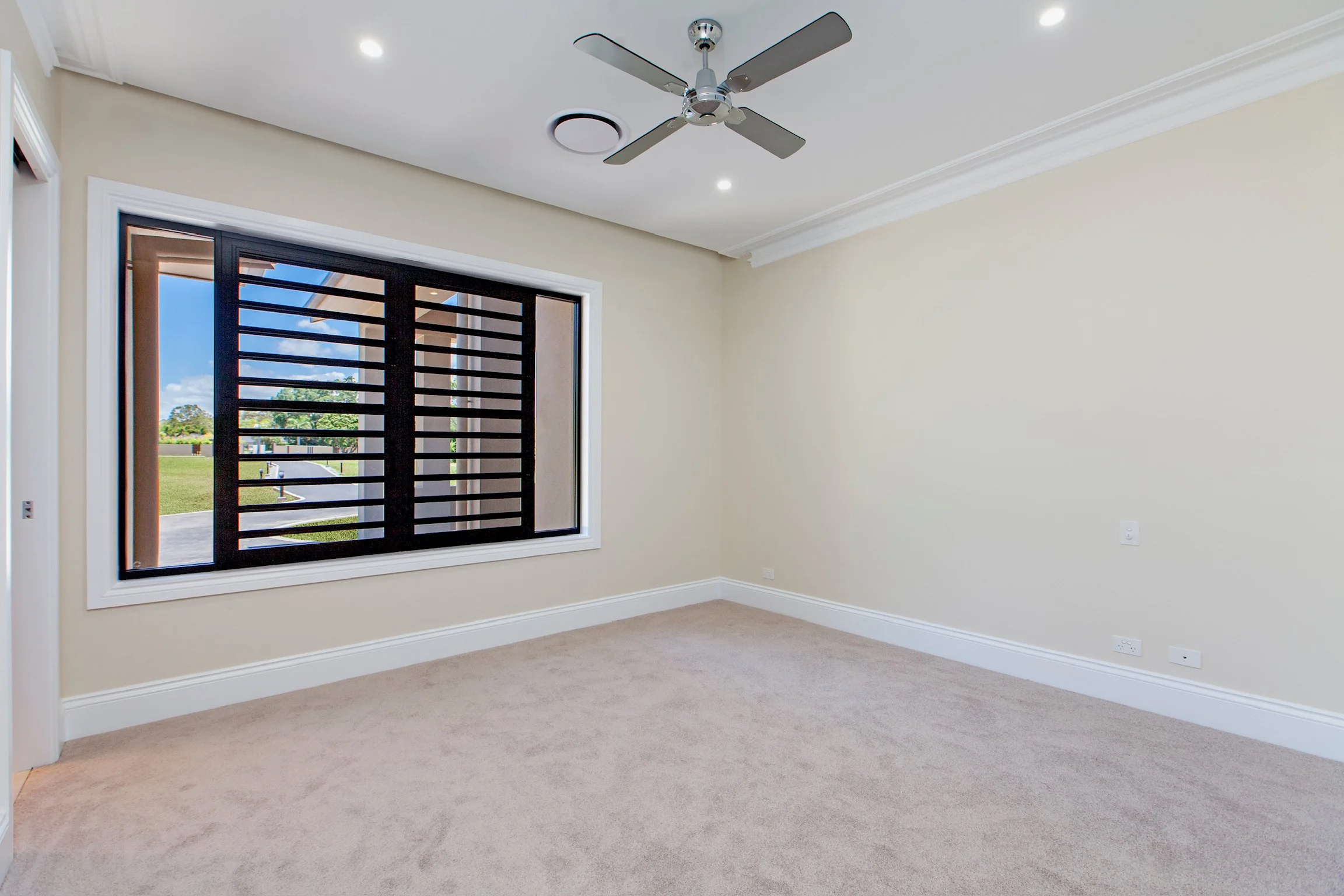 Empty room with beige carpet, white walls, a large window with black slatted shades, and a ceiling fan with five blades.
