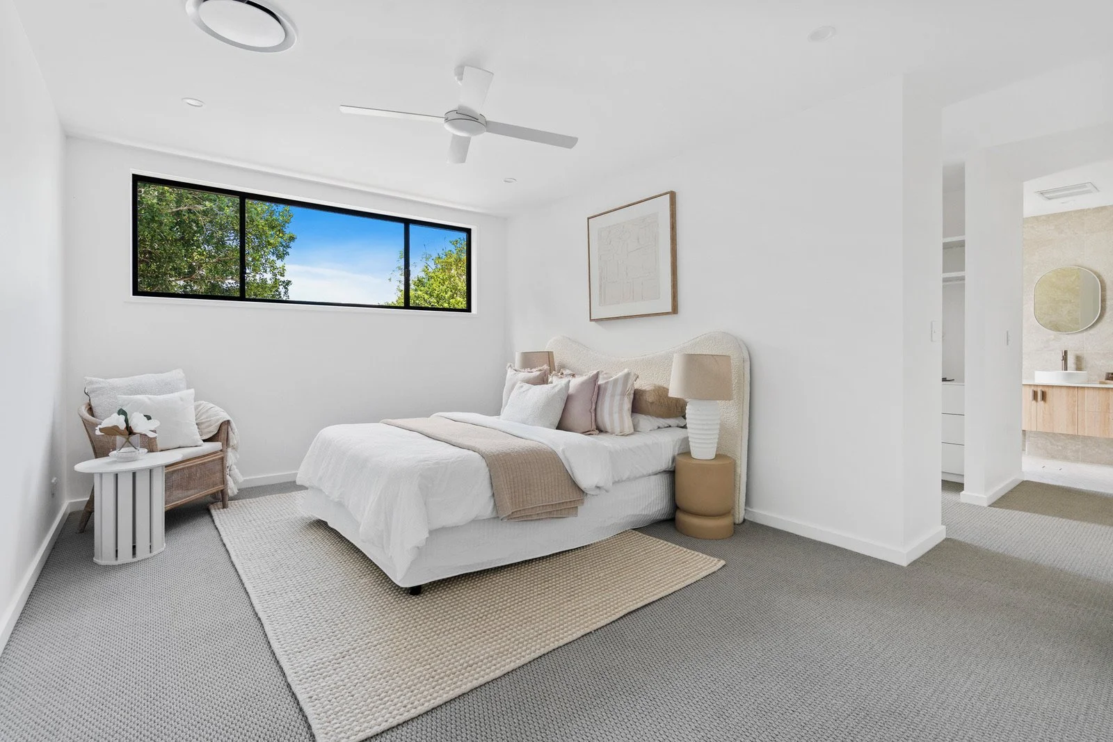 Minimalist white bedroom with a large window showing greenery outside, a bed with beige and white bedding, and simple decor including a side table with a lamp and a chair with pillows.