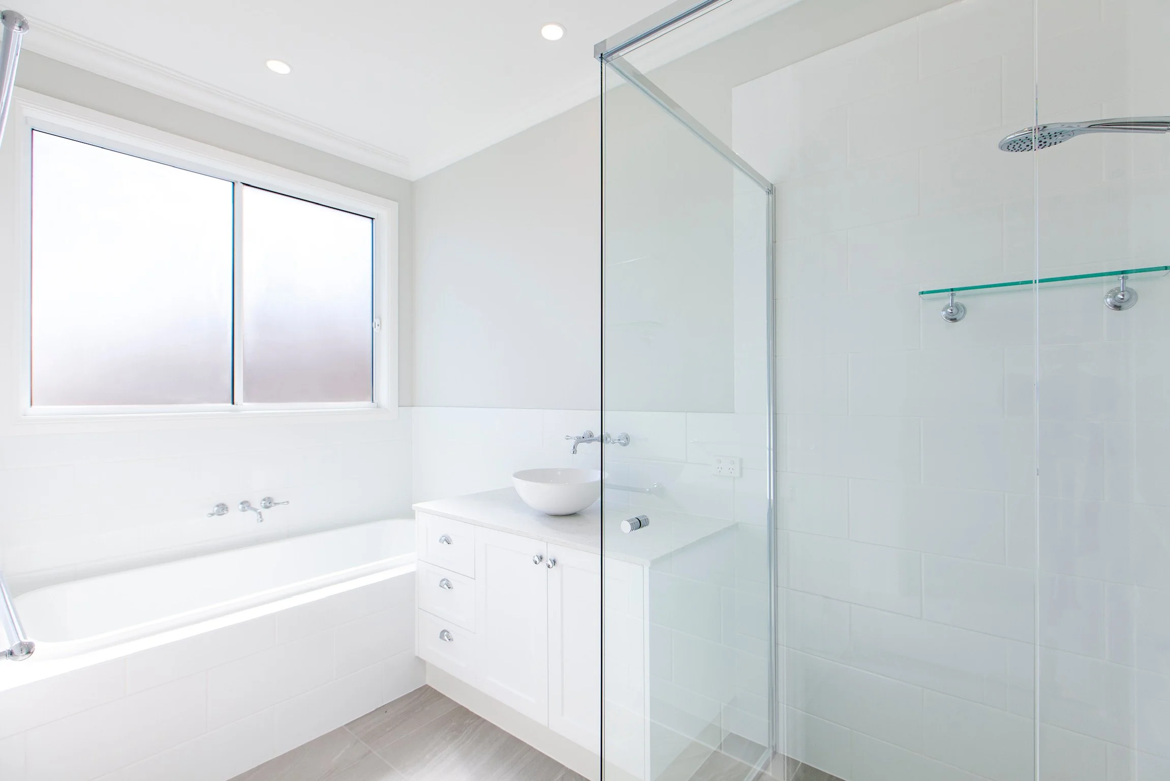 A modern, bright bathroom with white walls, a large frosted window, a glass shower enclosure, a white vanity with a bowl sink, and gray flooring.