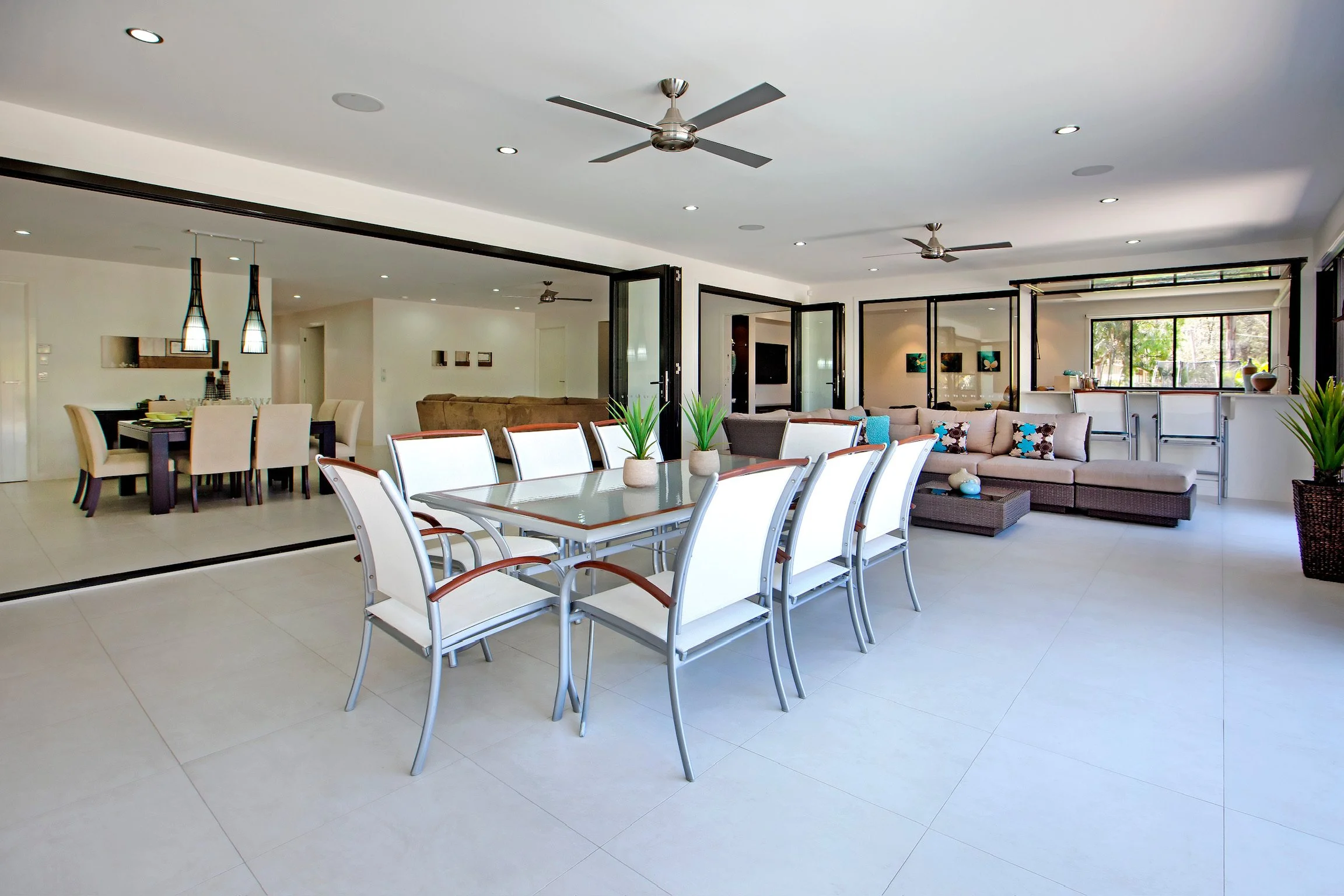Interior of a modern open-plan living and dining area with white walls, tiled flooring, ceiling fans, and large windows. The space features a dining table with white chairs, a sectional sofa with colorful cushions, and decorative plants.