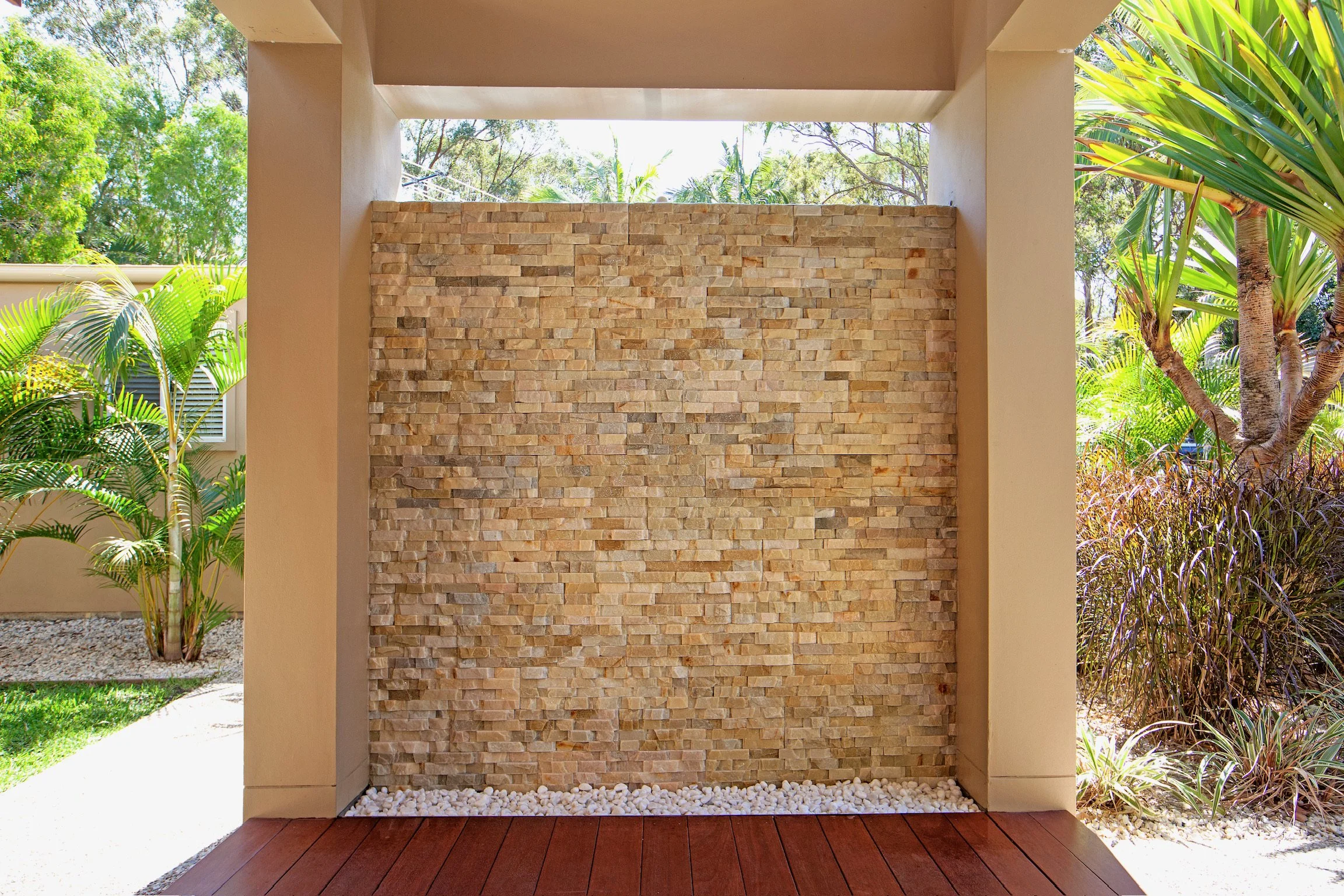 Outdoor wall with stacked stone design, framed by beige columns, with surrounding tropical plants and trees in the background.