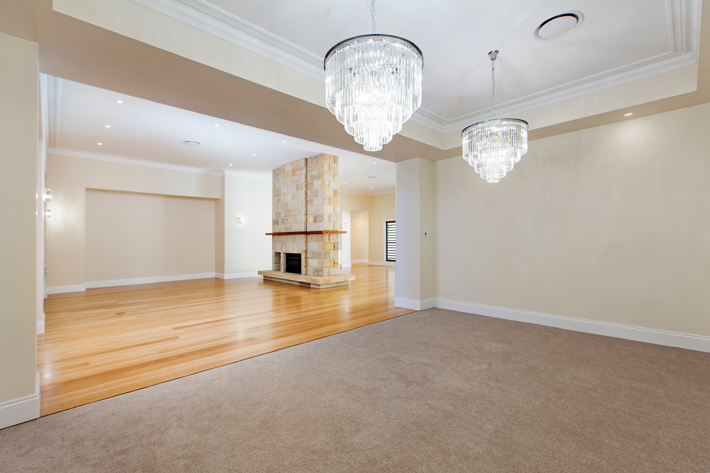 Empty living room with beige walls, hardwood flooring, a stone fireplace, and two modern chandeliers hanging from the ceiling.