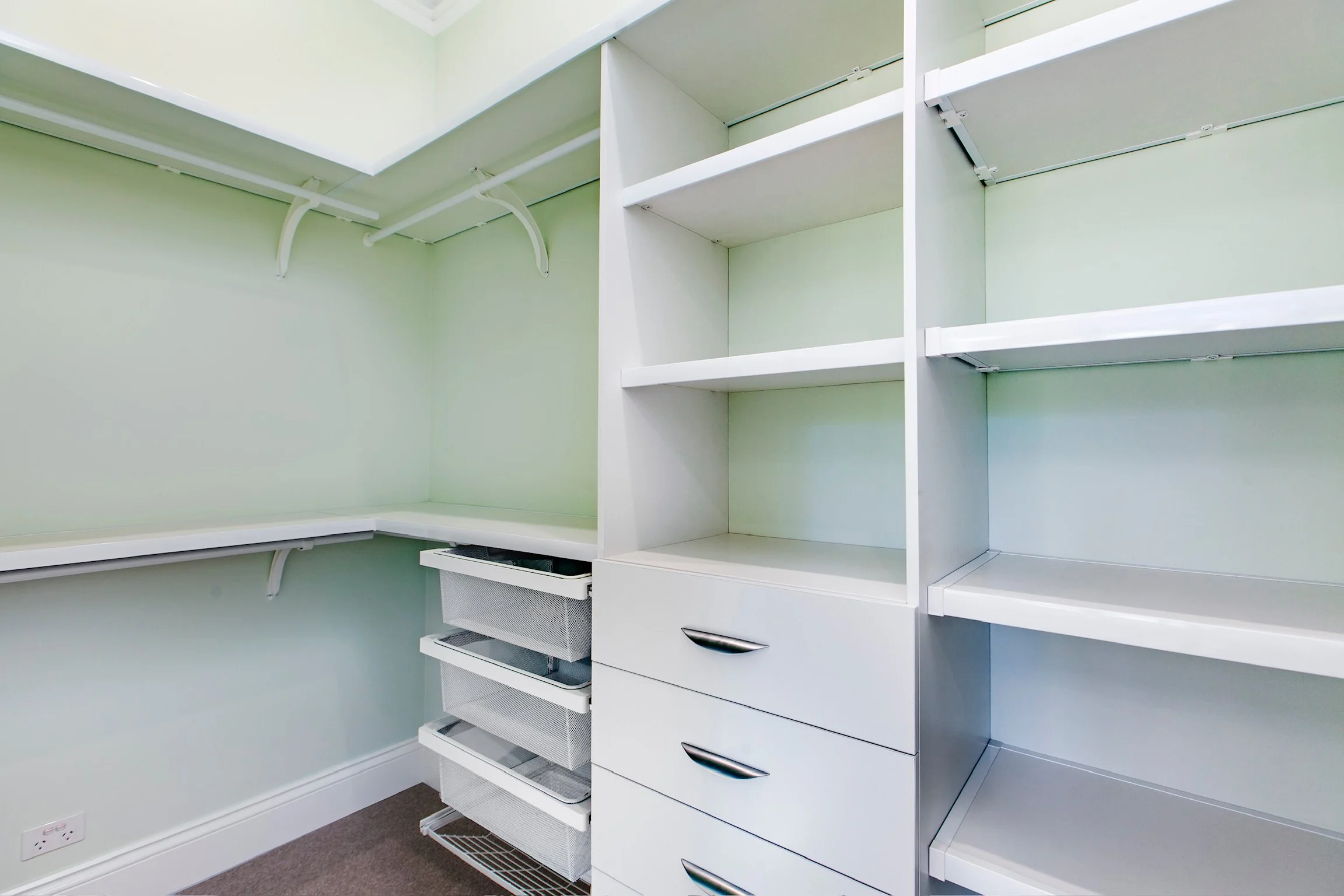 Empty white built-in closet with open shelving, drawers, and mesh baskets in a room with light green walls and beige carpet.
