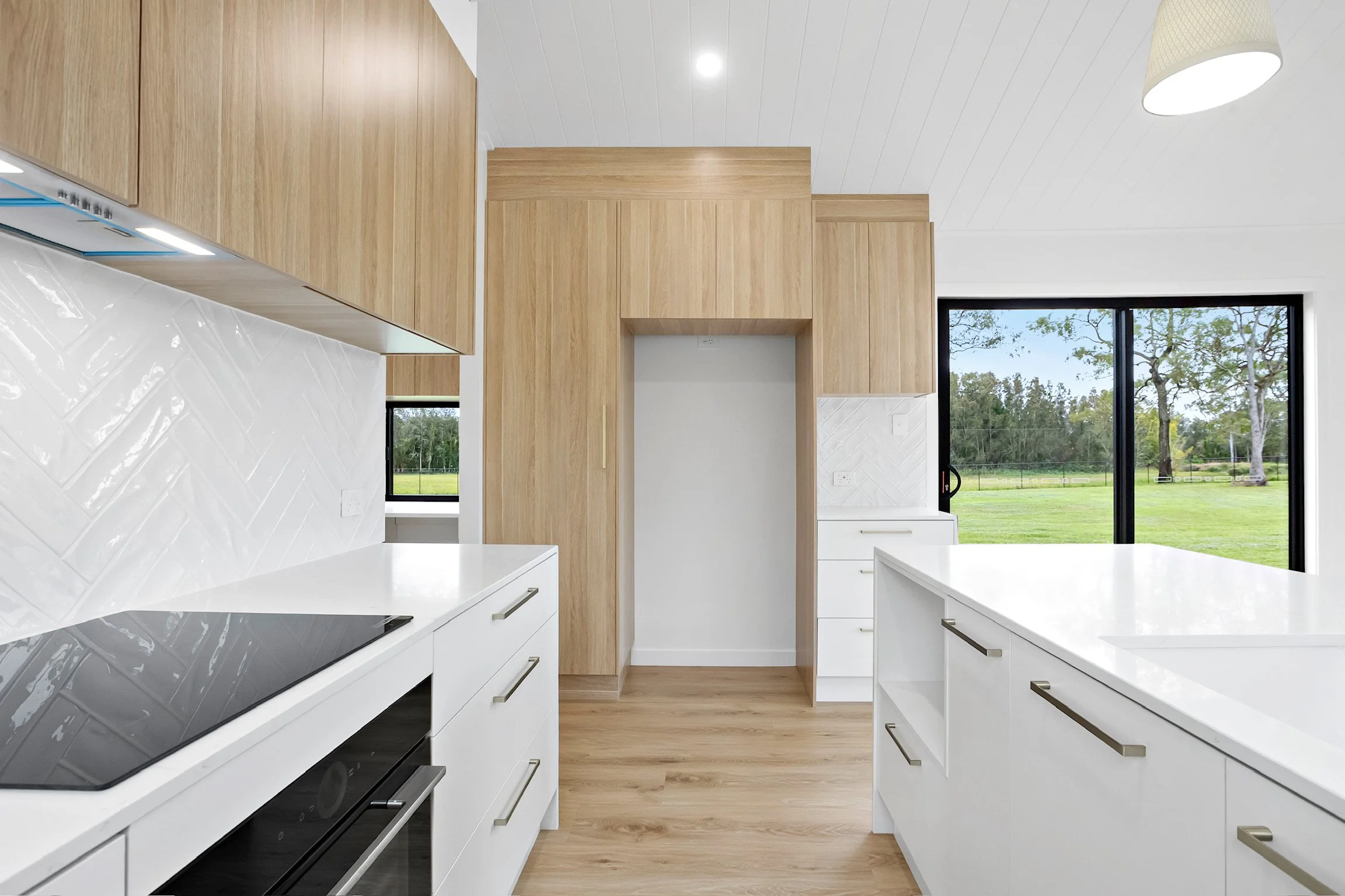 Modern kitchen with white cabinets, black countertop stove, wooden cabinets, large window overlooking trees, white tiled backsplash, and white ceiling with a light fixture.